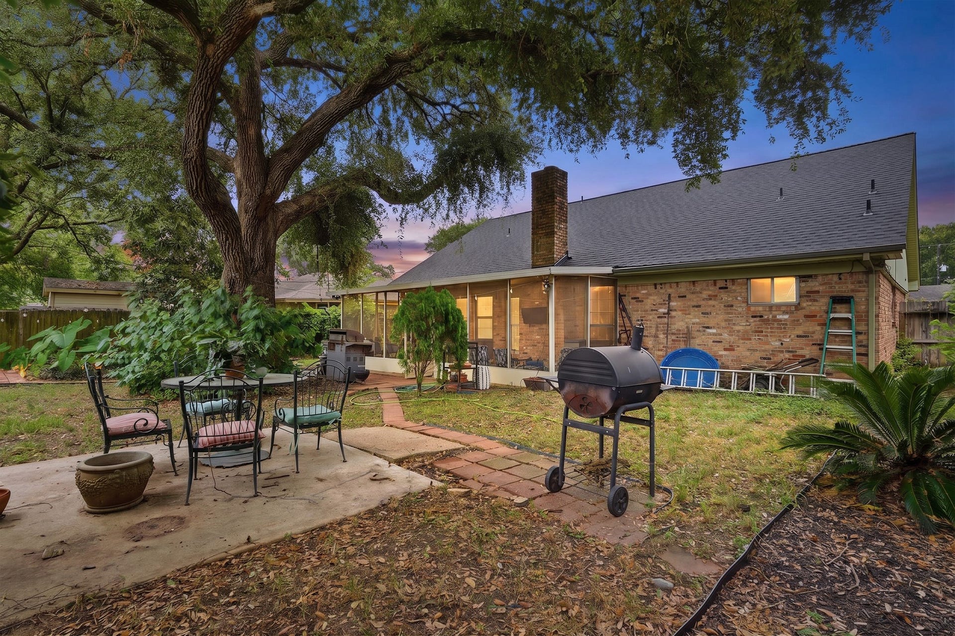 2216 Peach Lane, Pasadena, TX 77502 A cozy backyard with a charming brick house, featuring a large tree providing shade. Theres a patio area with outdoor seating and a grill, perfect for entertaining. The spacious yard is surrounded by greenery, offering a tranquil setting.