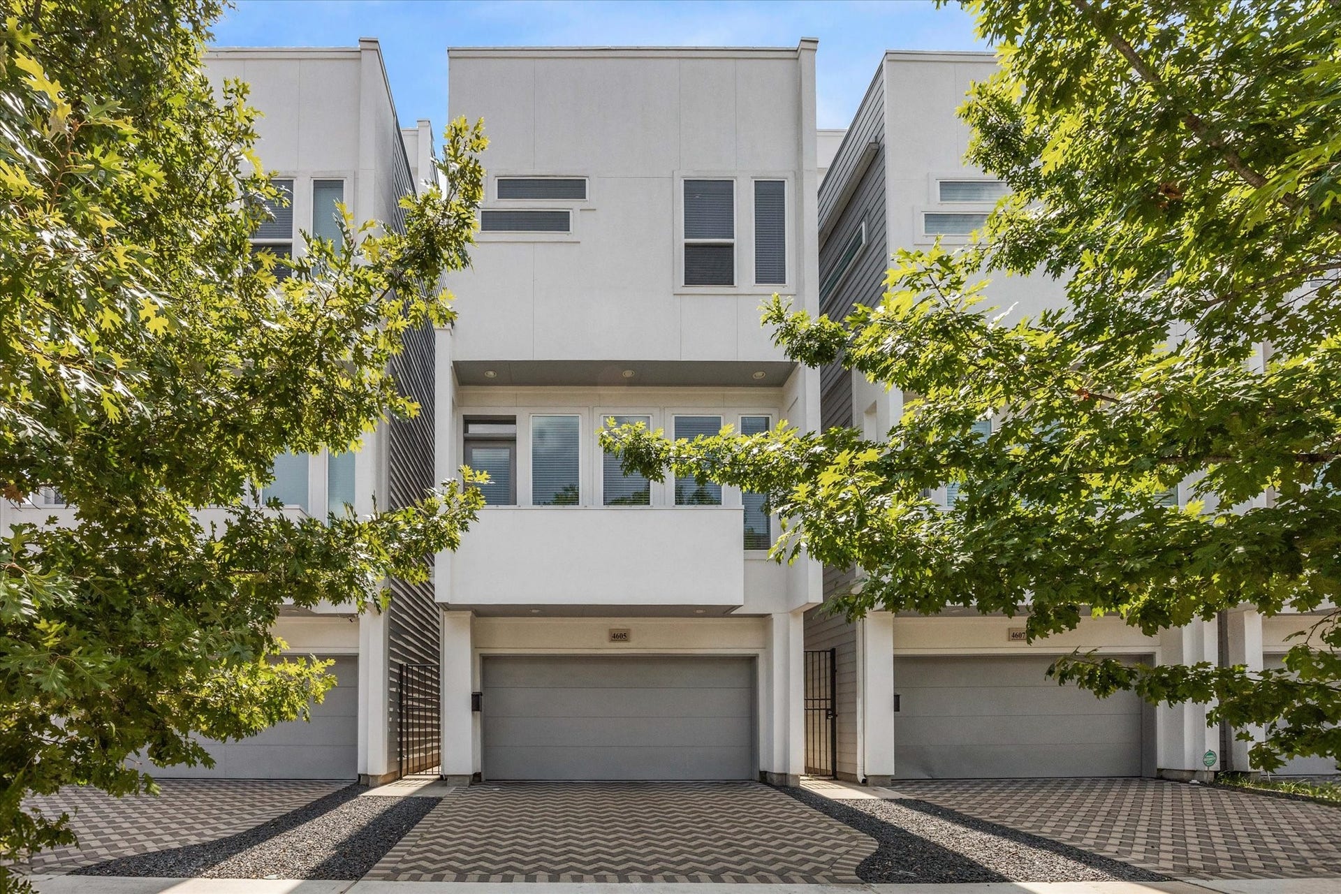 Contemporary three-story home framed by mature trees, featuring a clean stucco exterior, modern architectural lines, and an attached two-car garage. A patterned paver private driveway and landscaped approach add texture and curb appeal to the sleek urban design.
