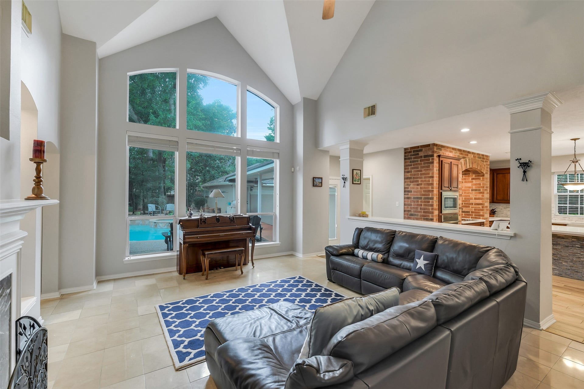 7 Larkspur Trail, The Woodlands, TX 77382 Spacious living area with soaring vaulted ceilings, large windows overlooking the pool, and a cozy fireplace, seamlessly connected to a modern kitchen with brick accents.