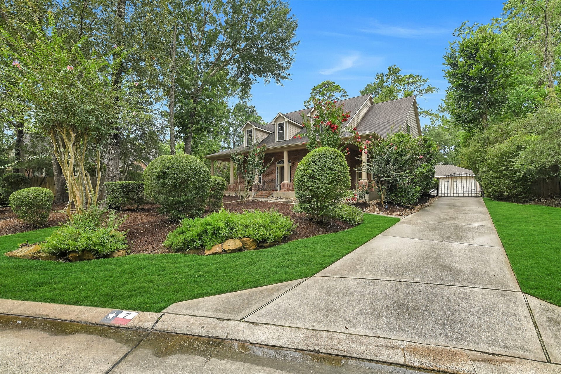 7 Larkspur Trail, The Woodlands, TX 77382 Charming home with a spacious front porch, surrounded by lush greenery and mature trees, featuring a long driveway leading to a detached garage.