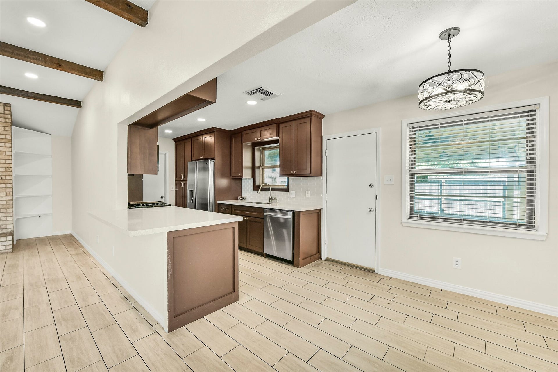 5003 Colombia Drive, Pasadena, TX 77505 Kitchen with sleek stainless steel appliances, rich wood cabinetry, and a spacious breakfast bar. The open layout flows into the living area, highlighted by exposed beams and a cozy brick accent wall with built-in shelving. Natural light pours in through the large window, enhancing the inviting atmosphere.
