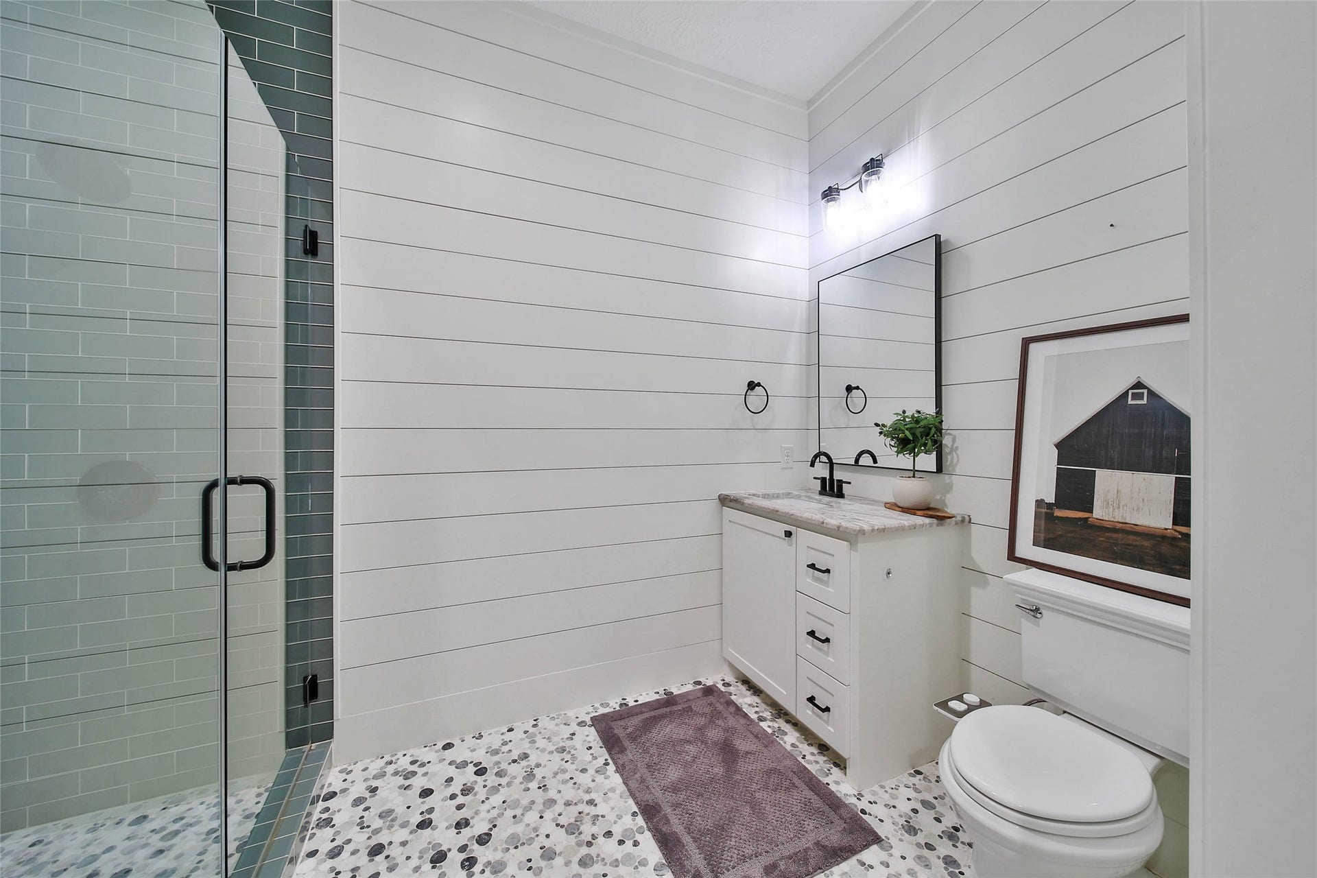 Designer full bath featuring a frameless glass walk-in shower with floor-to-ceiling tile, custom cabinetry, shiplap accent walls, pebble tile flooring, and stylish matte black fixtures.