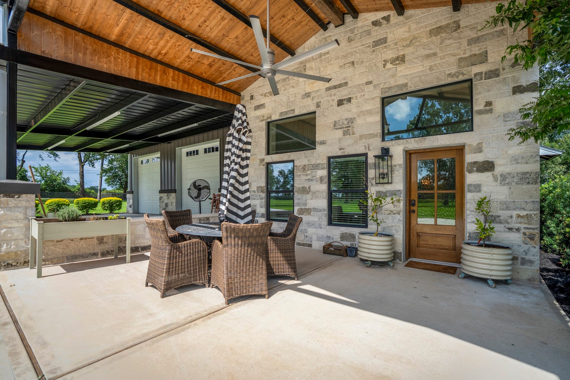 Covered patio outside the detached garage apartment showcasing tongue-and-groove wood ceiling, exposed steel beams, stone faade, oversized Big Ass Fan ($3000), and spacious outdoor seatingperfect for relaxing or entertaining year-round.