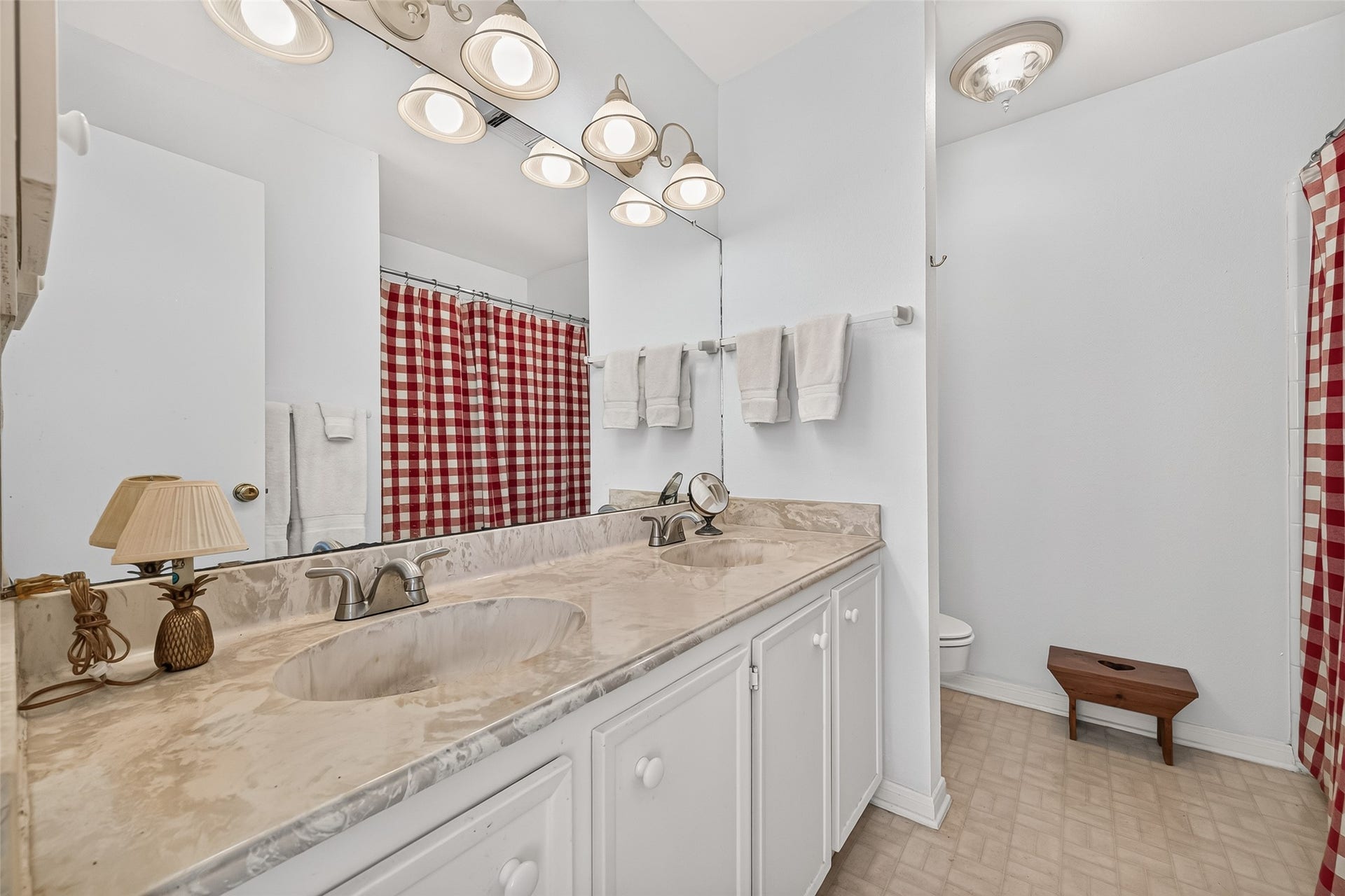 Bright and spacious secondary full bathroom featuring a double vanity with elegant marble countertops, classic white cabinetry, and charming red checkered shower curtains for a cozy touch.