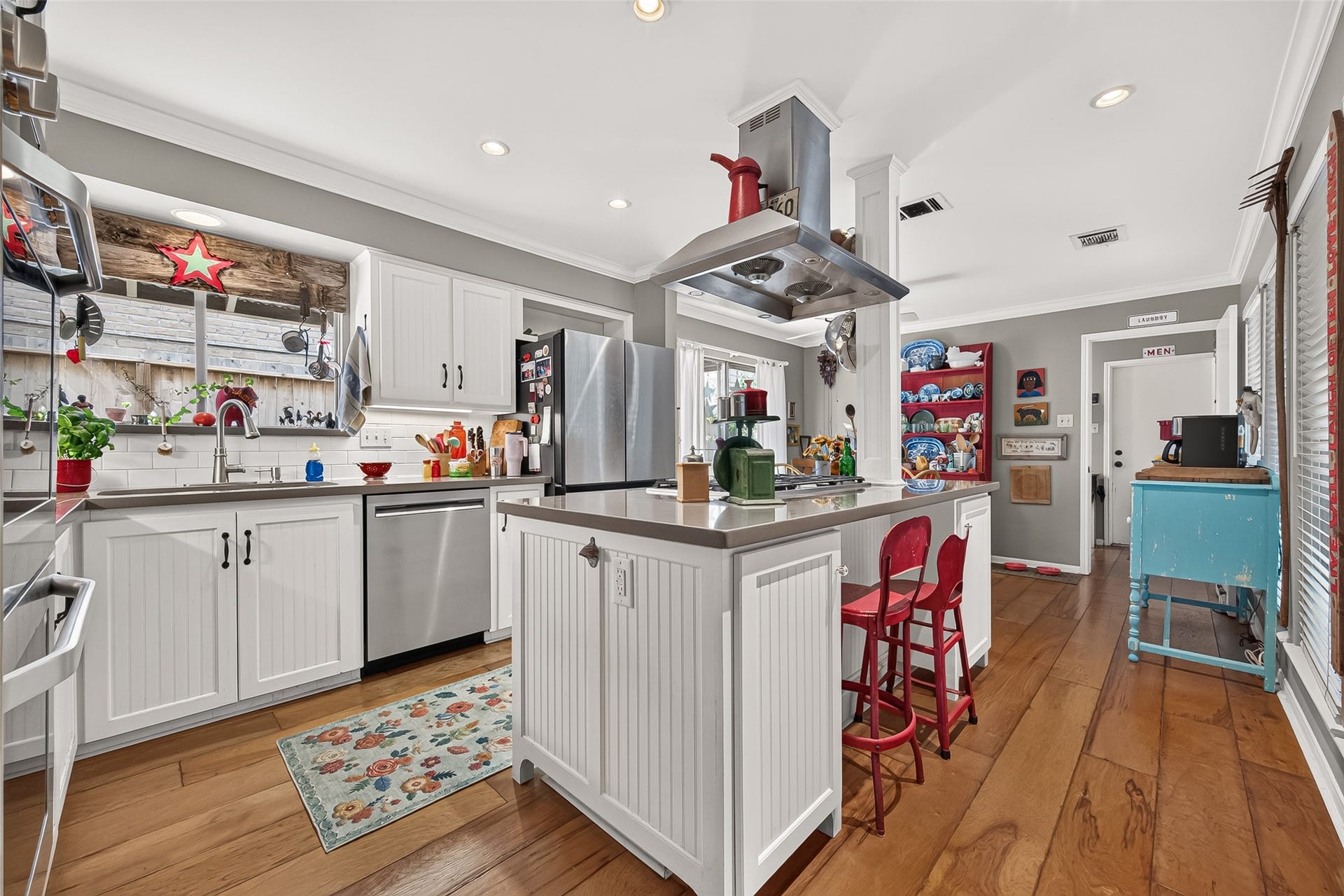 Kitchen with a modern farmhouse vibe, featuring white cabinetry, stainless steel appliances, and a central island with red bar stools. Natural light floods in through a large window, highlighting the warm wood flooring and colorful accents.