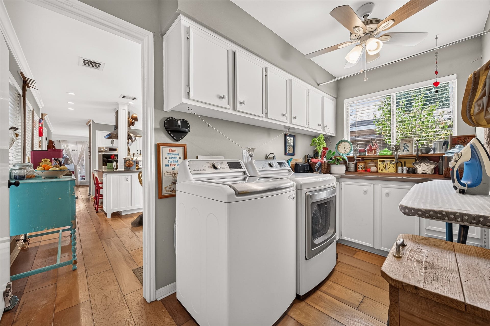 Laundry room featuring ample cabinetry, modern appliances, and a ceiling fan, seamlessly connected to an open kitchen area with warm wood flooring.