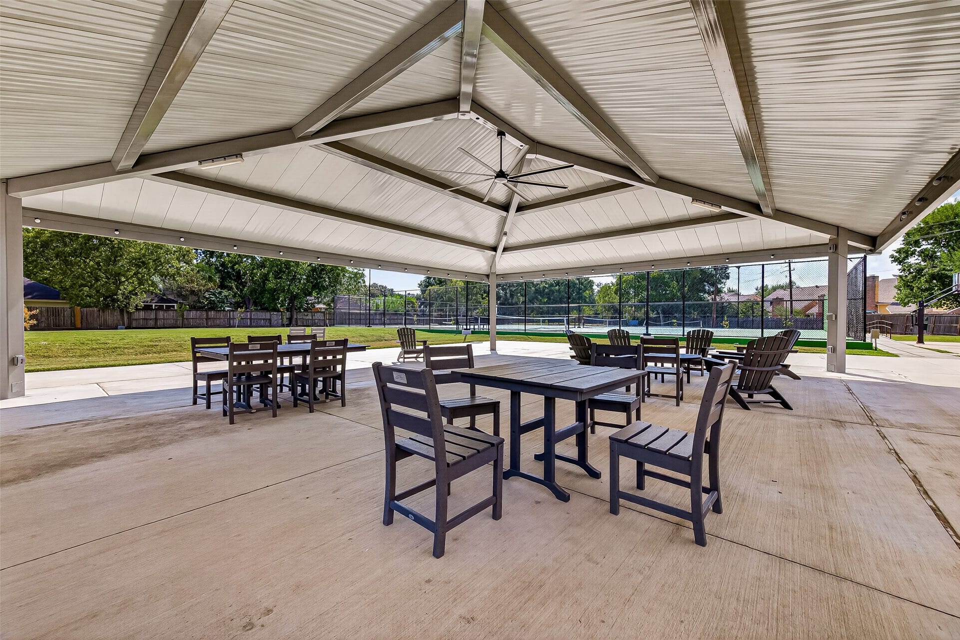 Spacious covered pavilion with ample seating, perfect for outdoor gatherings and relaxation, overlooking a lush green lawn and tennis courts.