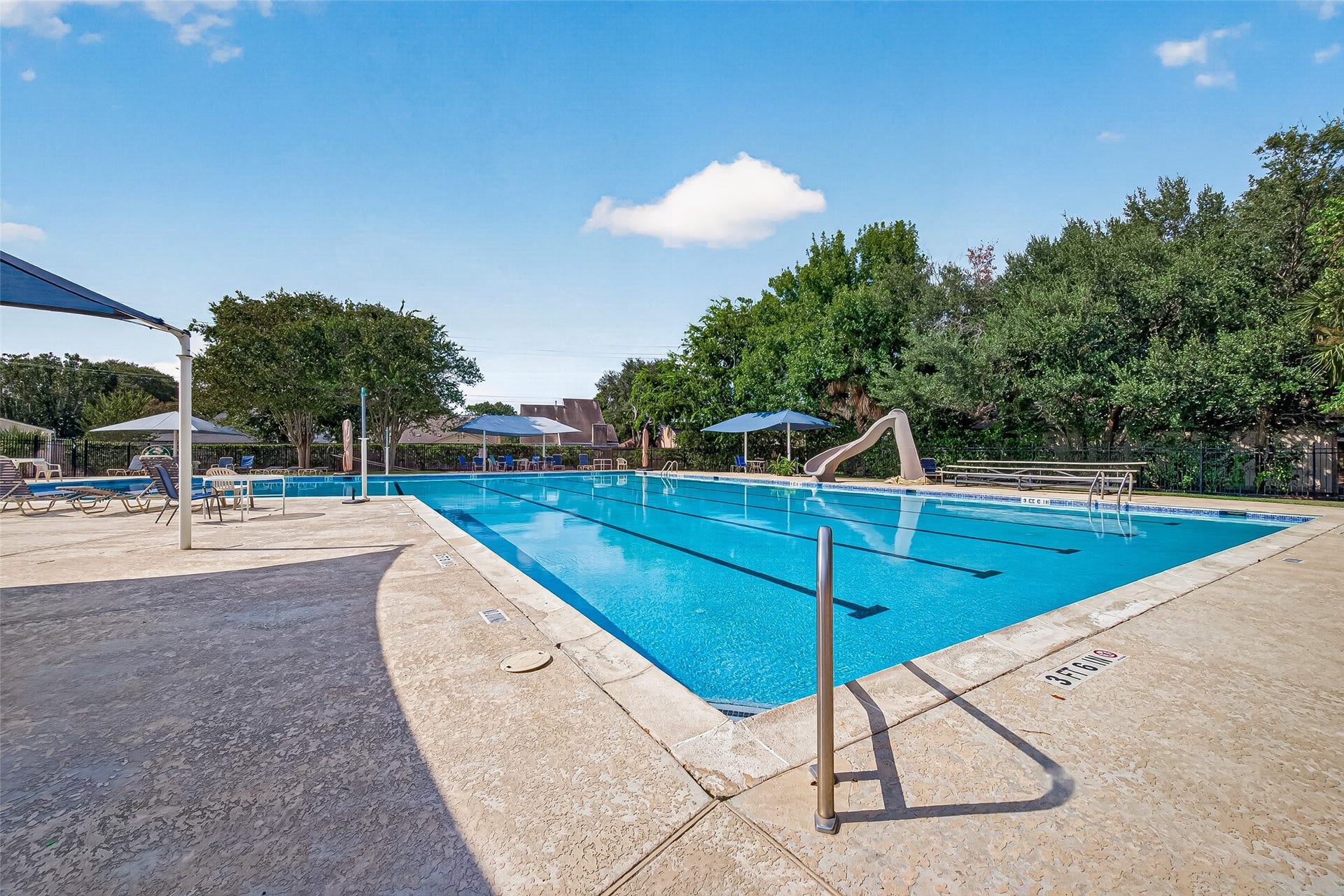 Enjoy a refreshing swim in this spacious community pool, complete with a fun slide and ample lounging areas under shaded umbrellas, nestled amidst lush greenery.