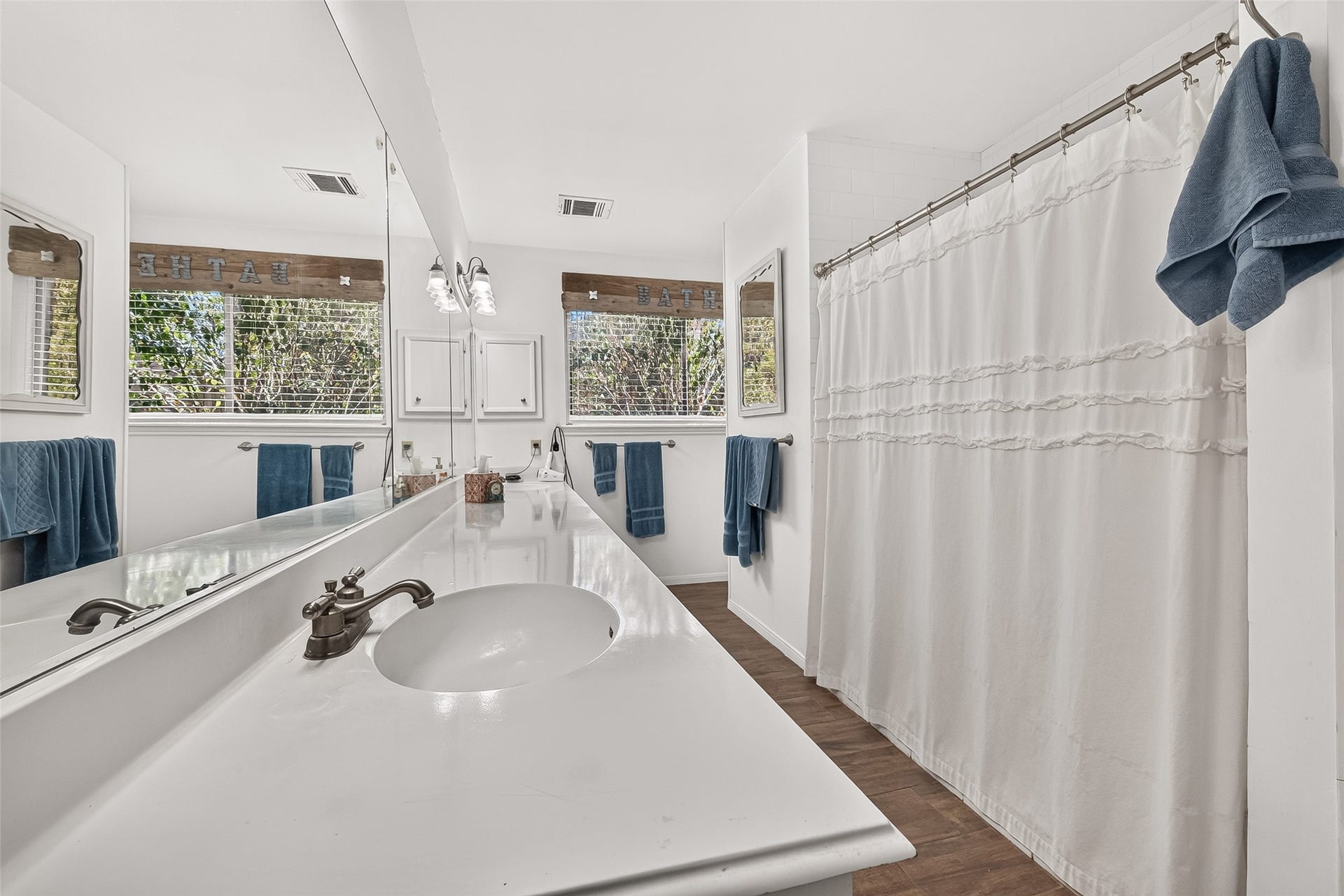 Bright and inviting primary bathroom featuring a long vanity with dual sinks, a spacious mirror, and a shower with elegant ruffled curtains. Natural light flows in through the window, complementing the clean, modern design.