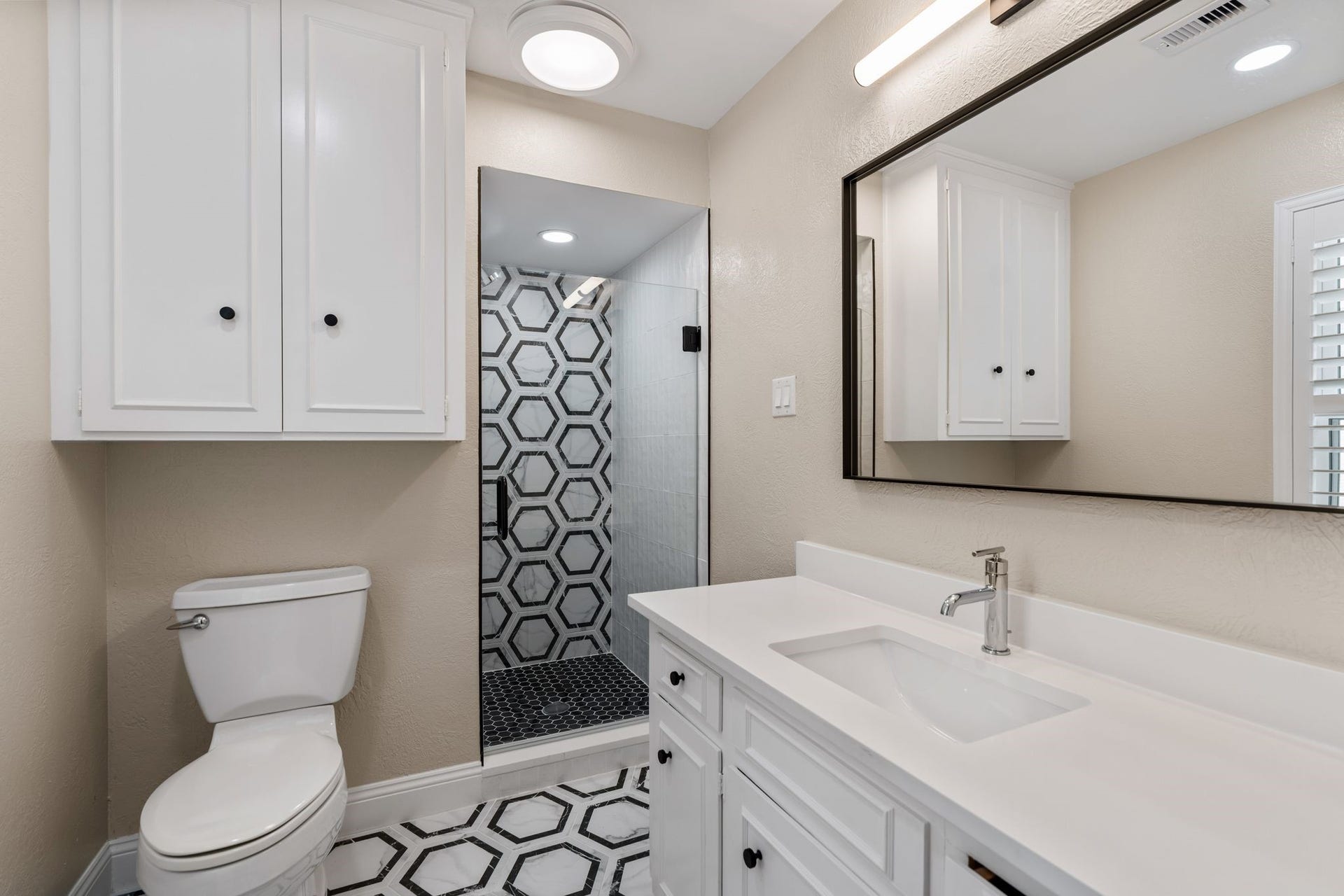 8619 La Fonte Street, Houston, TX 77024 The ensuite bathroom features black-and-white geometric tile flooring, a vanity with quartz countertops and cabinet and drawer storage, and a decorative matte black mirror with coordinating lighting. The walk-in shower is finished with a glass door, black hexagon tile flooring, a decorative accent wall and white subway tile on the surrounding walls. Additional cabinetry above the toilet provides extra storage.
