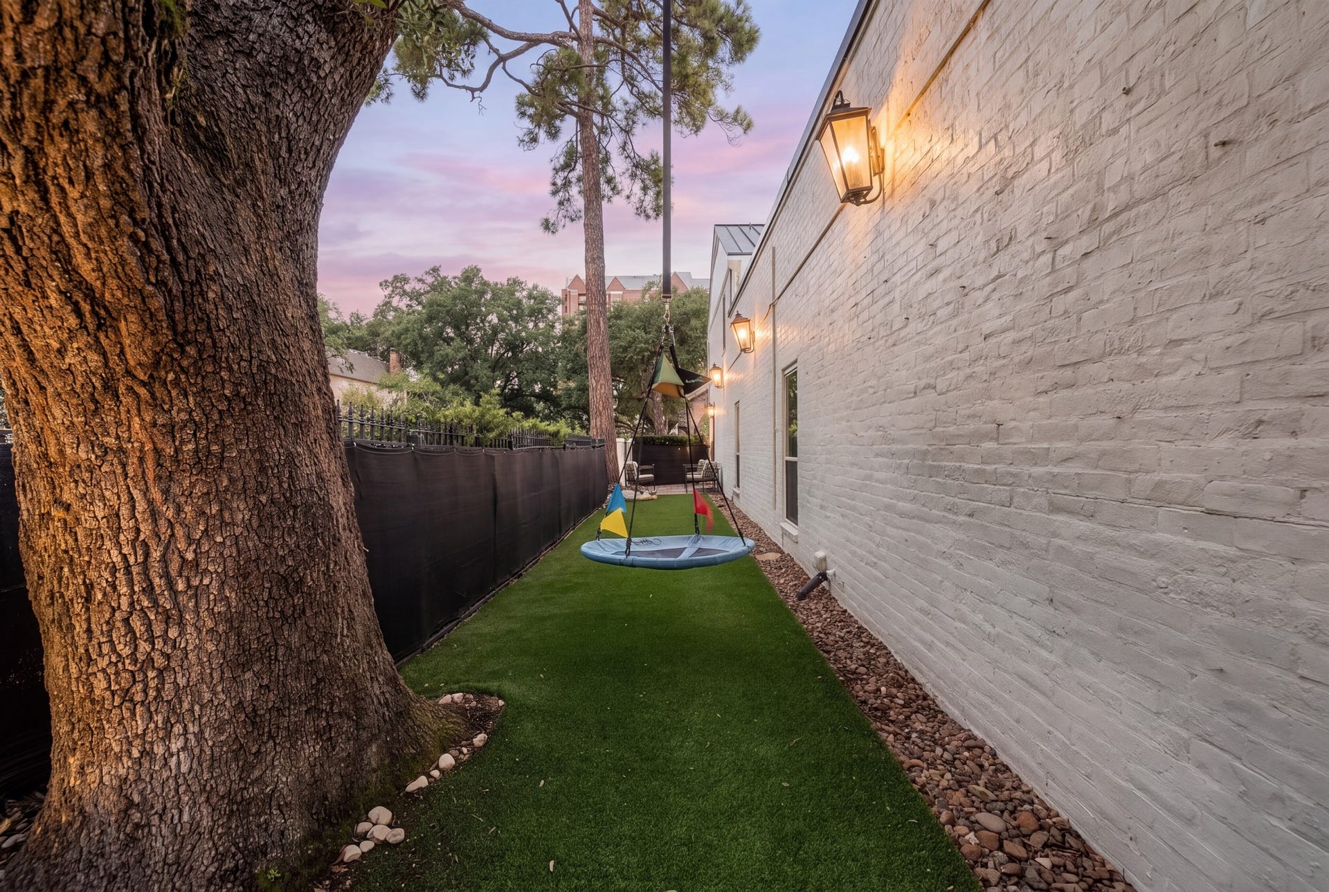 8619 La Fonte Street, Houston, TX 77024 Shown at dusk, the side yard is enhanced by exterior lantern lighting along the white brick wall, creating a warm evening setting. The combination of turf, mature landscaping and usable open space makes this an especially functional outdoor area.