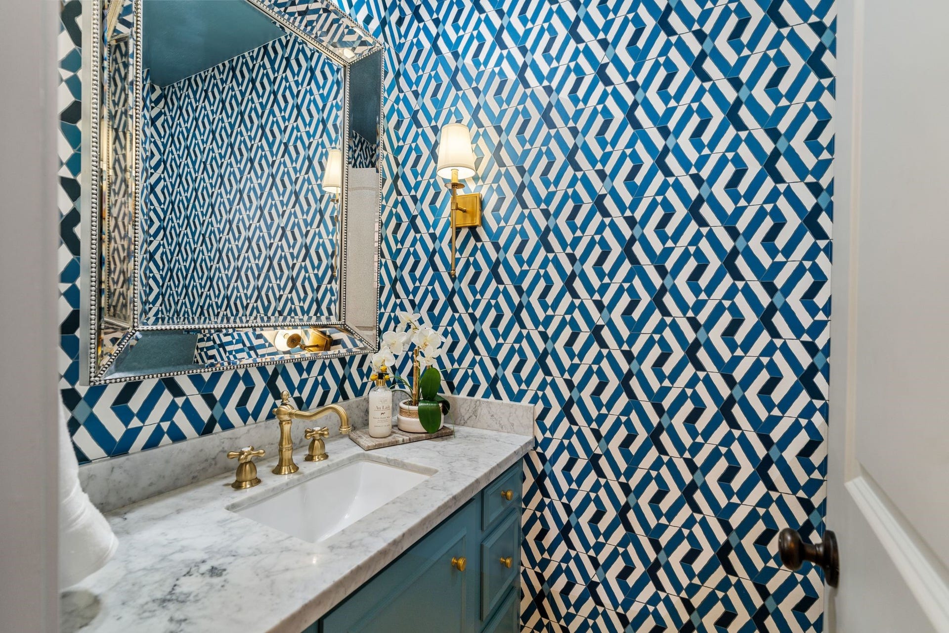 8619 La Fonte Street, Houston, TX 77024 The powder room makes a bold statement with decorative blue-and-white wallpaper, a built-in vanity with accent-colored cabinetry, marble countertops and gold plumbing fixtures. Gold sconces flank the decorative mirror, while the window with shutters brings in natural light. Tucked away from the main living areas, the powder room offers added privacy while still feeling polished and thoughtfully finished.