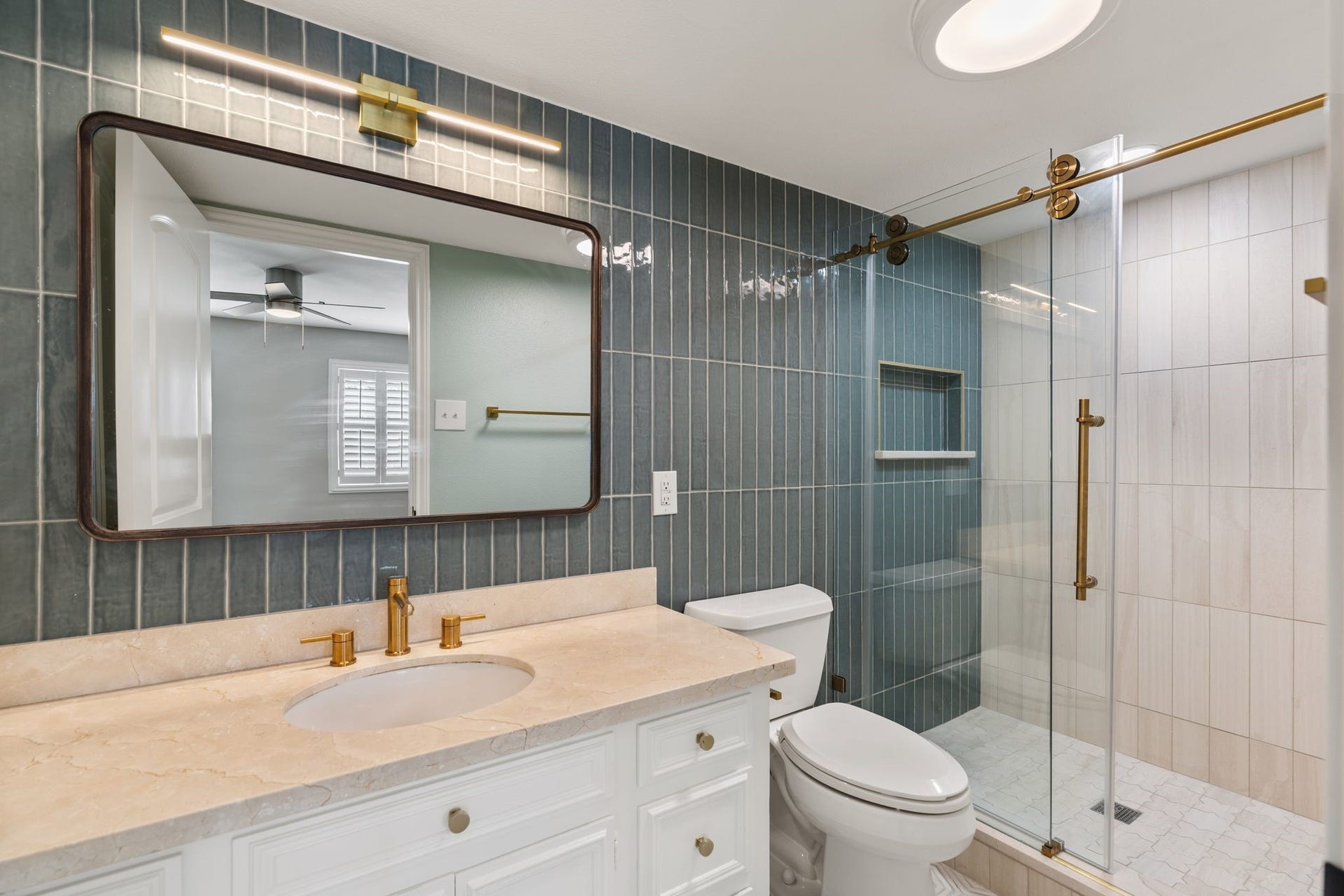 8619 La Fonte Street, Houston, TX 77024 This ensuite bathroom features a floor-to-ceiling blue tile accent wall, a white vanity with stone countertops and built-in storage, and a large framed mirror with coordinating gold vanity lighting. The walk-in shower includes a sliding glass door, brass-toned fixtures and a built-in niche, while additional cabinetry beside the vanity provides even more storage.