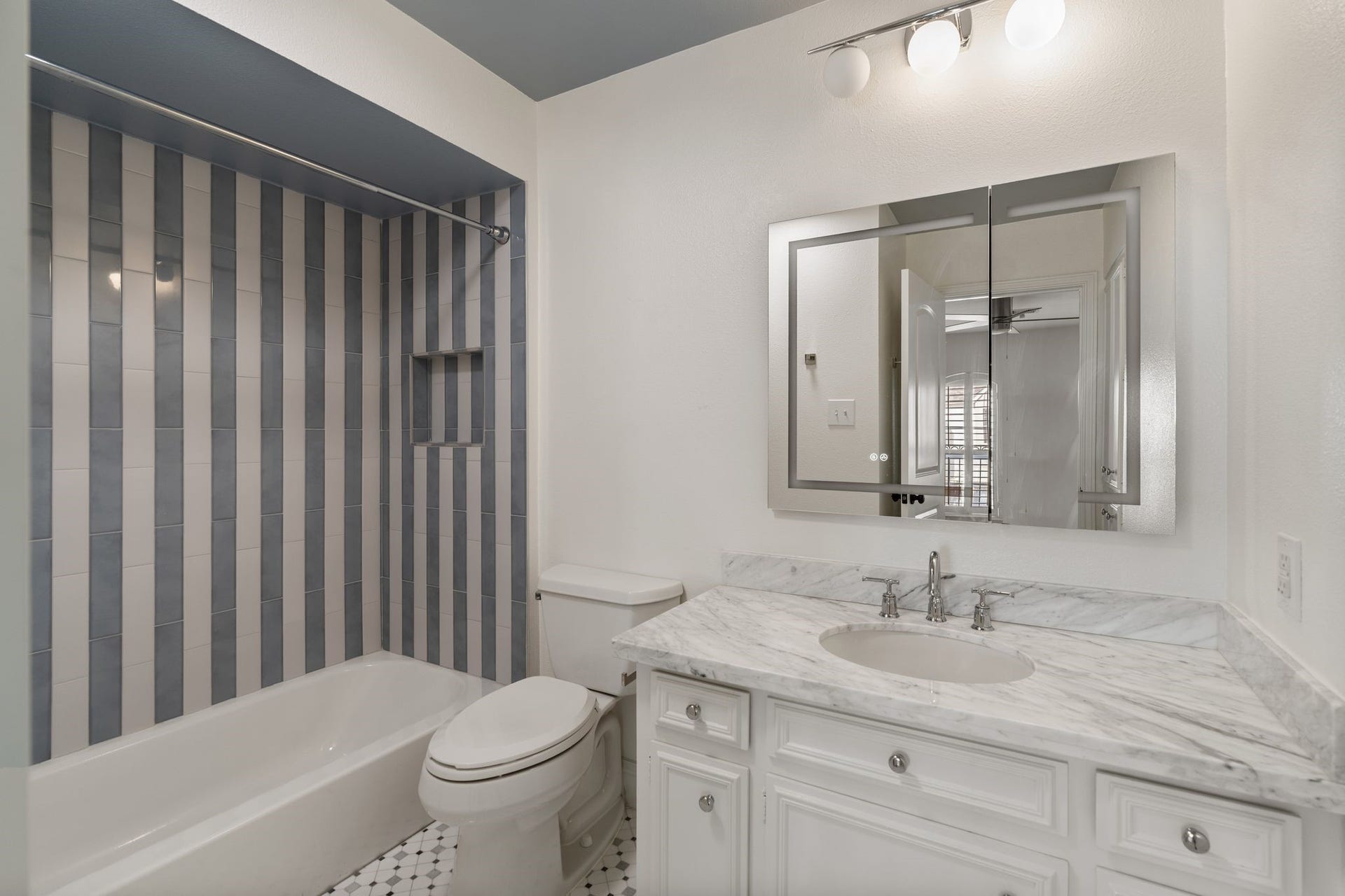 8619 La Fonte Street, Houston, TX 77024 The ensuite bathroom features decorative tile flooring, a white vanity with marble countertops and storage, a lit medicine cabinet mirror and additional built-in linen cabinetry. The tub/shower combination is finished with a striped subway tile surround and built-in shower niche, adding a polished touch to the space.