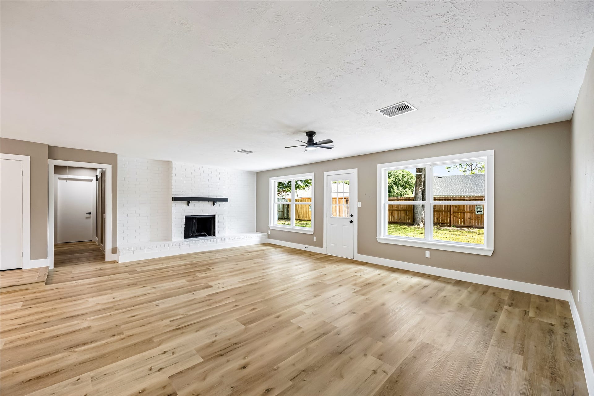 1803 Thornbrook Drive, Missouri City, TX 77489 Spacious living area featuring warm wood-look flooring, a white brick fireplace, modern ceiling fan, and large windows overlooking the backyard. The wide-open layout offers plenty of room for flexible furniture arrangements, while natural light creates a bright and welcoming feel.