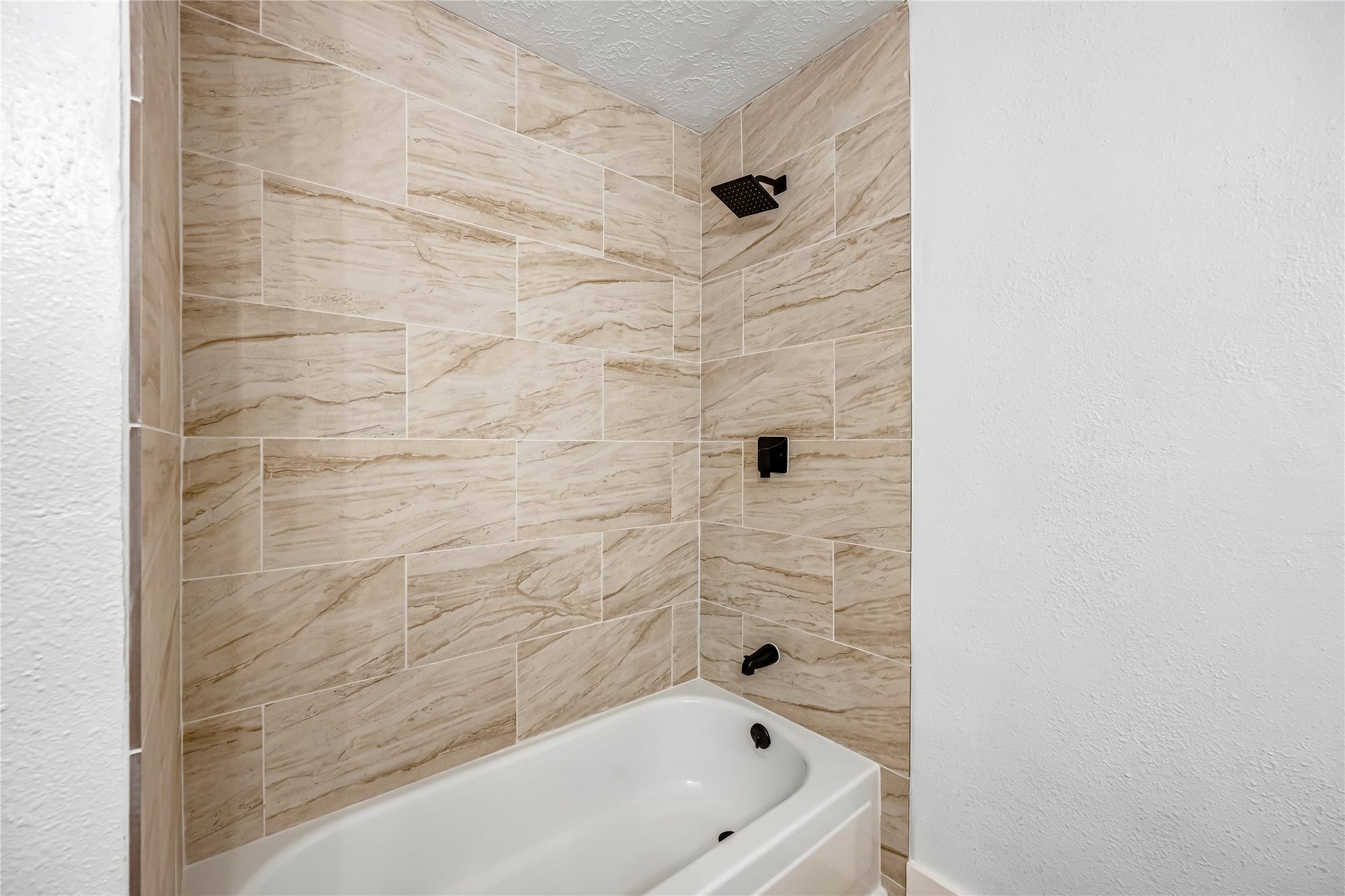 1803 Thornbrook Drive, Missouri City, TX 77489 Updated tub and shower area featuring warm marble-look tile, a clean white bathtub, and modern matte black fixtures. The floor-to-ceiling tile surround creates a polished, contemporary look with a warm and inviting finish.