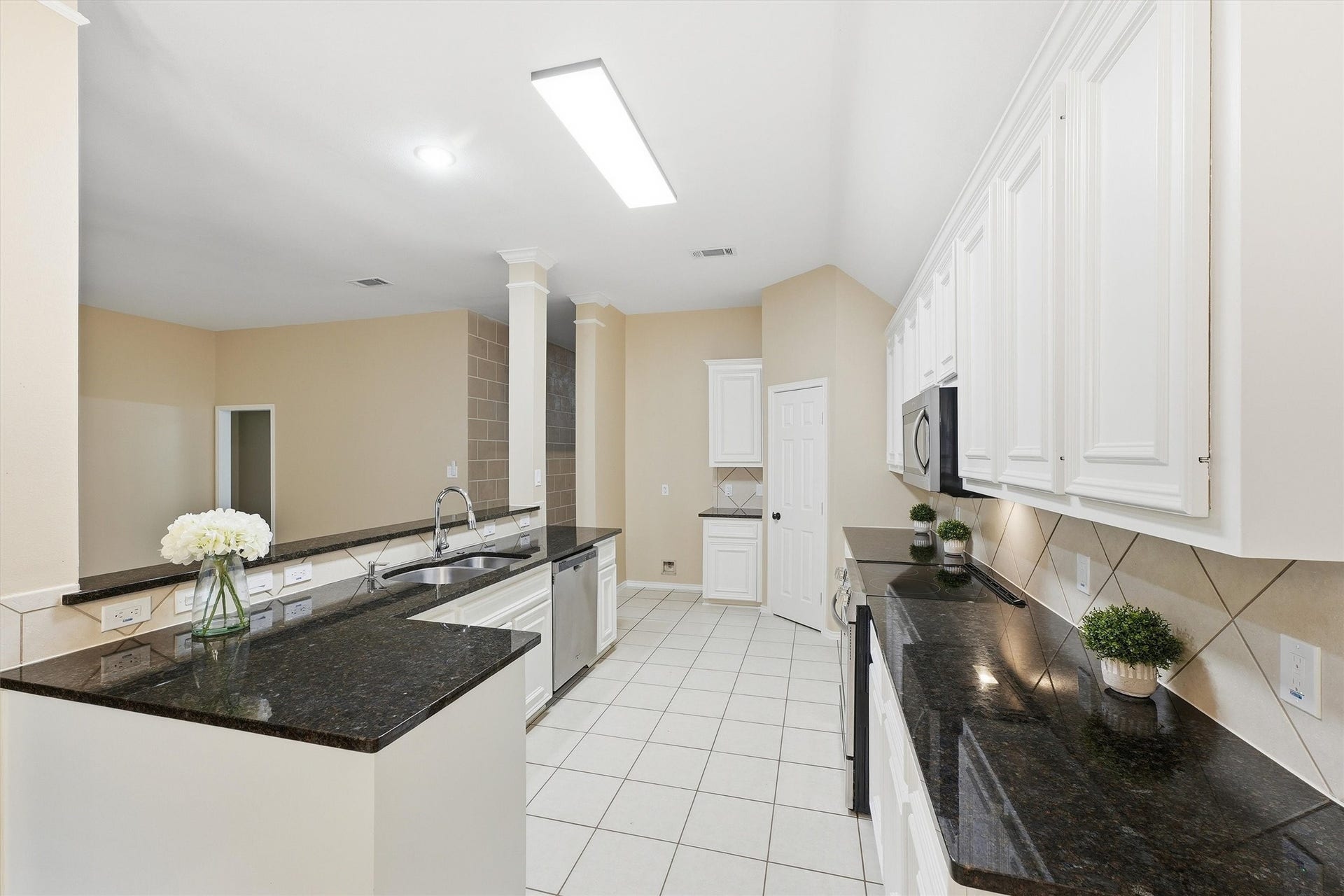 Galley-style kitchen equipped with dark granite countertops, crisp white cabinetry, tile floors, and a double stainless-steel sink.