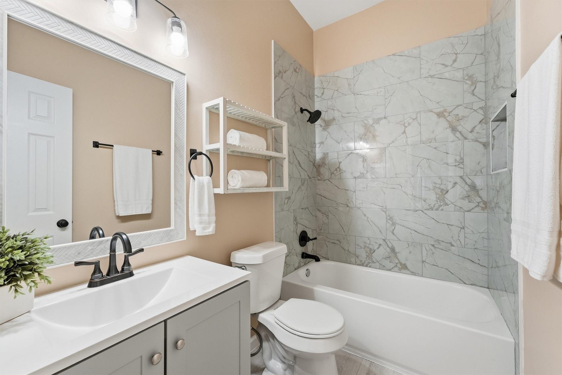 Updated guest bath features a modern grey vanity, matching framed mirror, oil-rubbed bronze fixtures, and a stunning marble-look tile shower surround.
