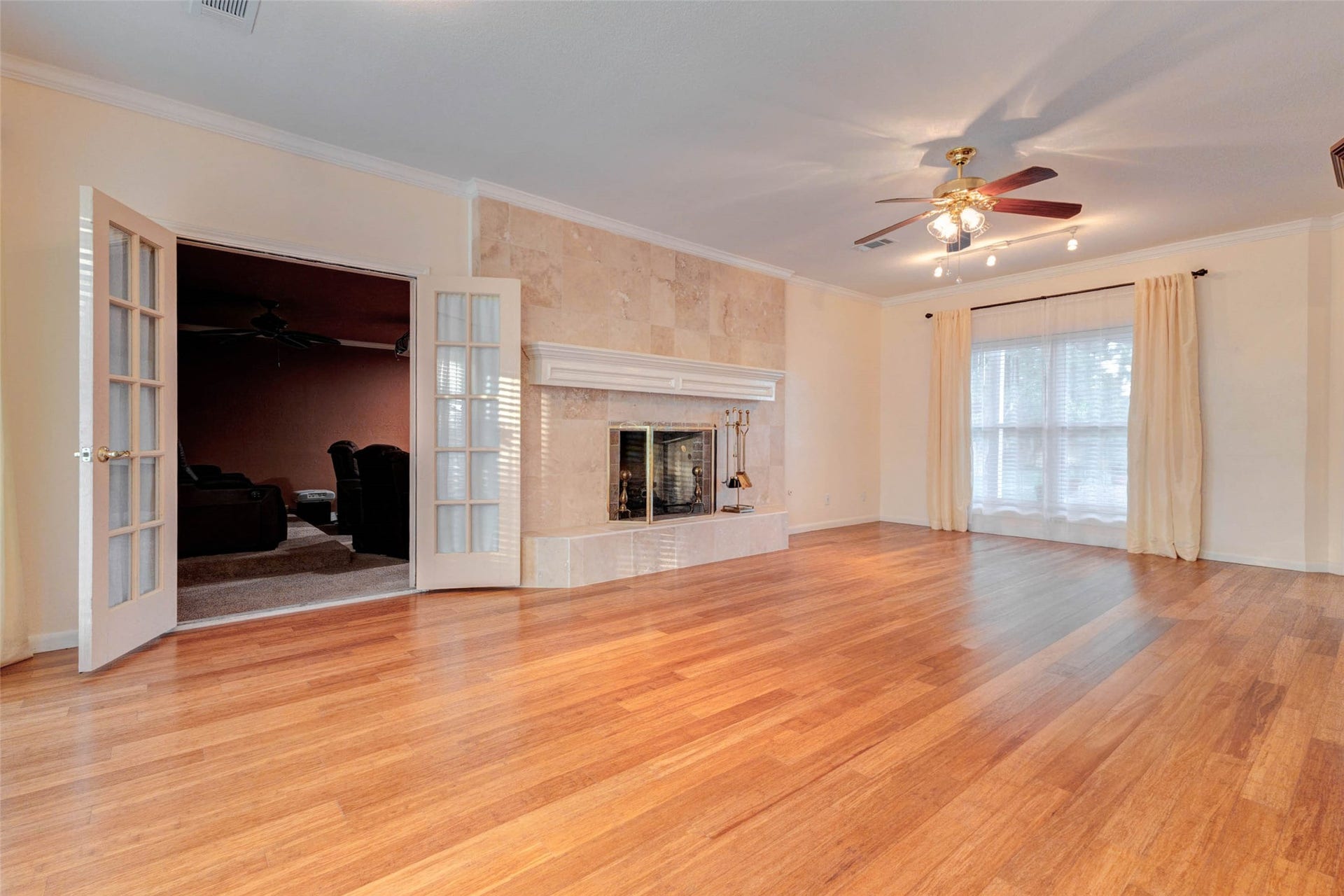 11418 Cedarwood Drive, Humble, TX 77338 This inviting living room offers generous open space, warm wood-look floors, crown molding, a ceiling fan, and a fireplace framed by neutral tile. French doors connect to an adjoining media or flex room, creating a versatile layout for entertaining, relaxing, or setting up multiple living zones within the home.