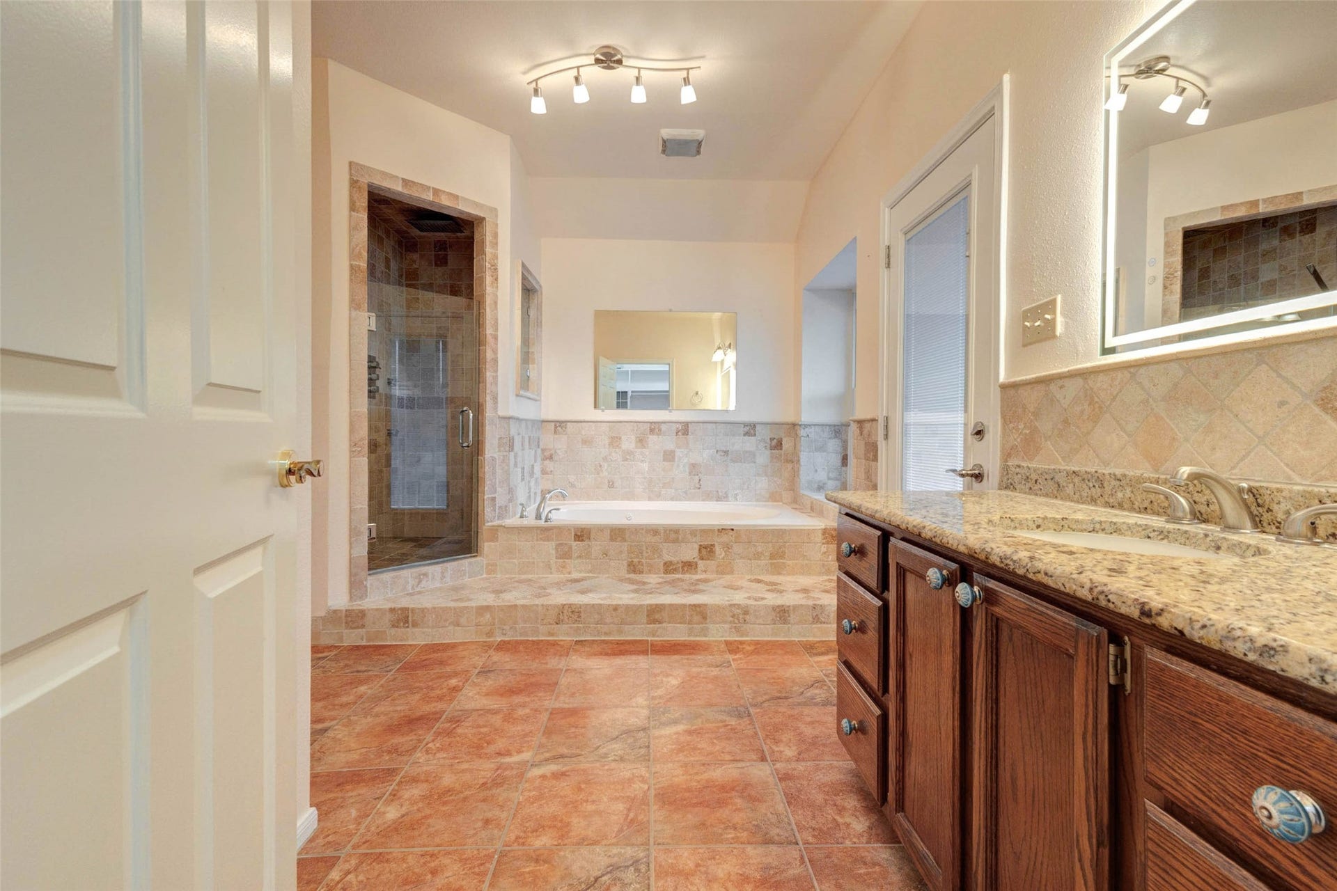 11418 Cedarwood Drive, Humble, TX 77338 This primary bath view highlights the separate glass-enclosed shower, jetted tub, tiled surround, granite-style vanity counter, warm cabinetry, and spacious floor plan. The layout offers both function and comfort, with multiple lighting points, neutral finishes, and a private bath design suited for everyday routines.