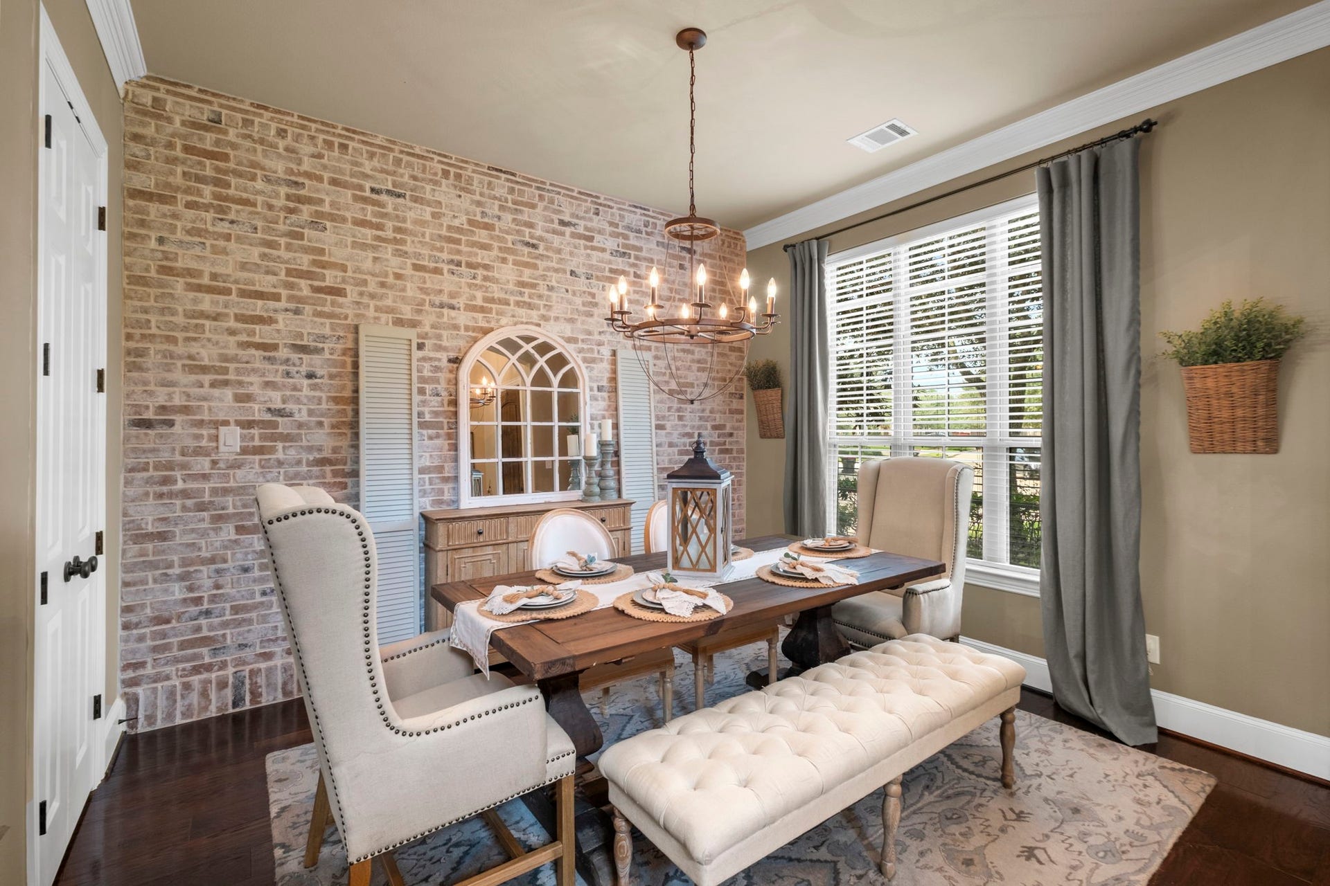 34 Stefano Way, Missouri City, TX 77459 Formal dining area highlighting an accent wall made of exposed brick finish, detailed crown molding, rich hardwood floors, and a wrought-iron candle-style chandelier. Large windows with custom blinds and drapery fill the space with natural light.