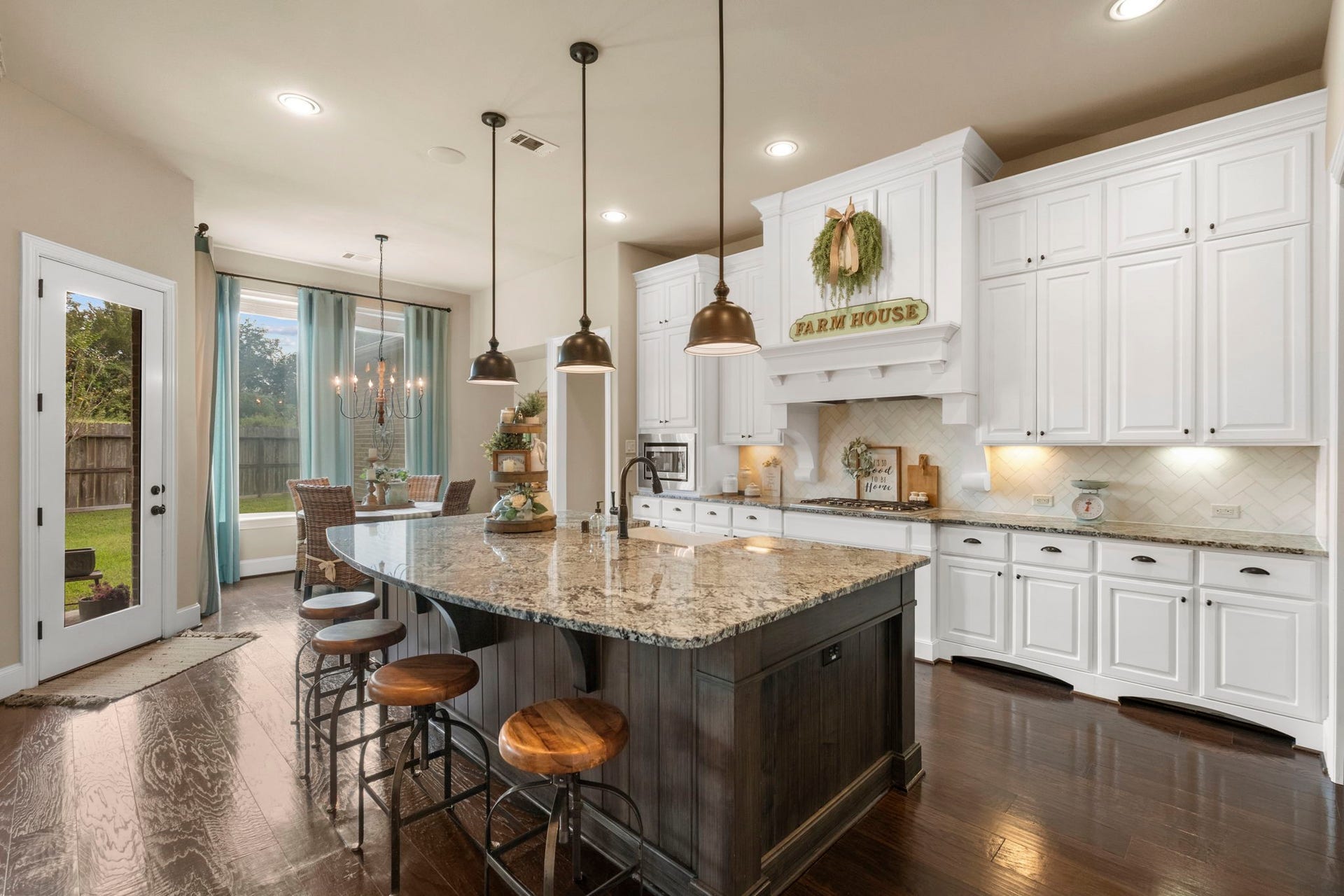 34 Stefano Way, Missouri City, TX 77459 Gourmet kitchen showcasing custom white cabinetry, dark hardwood floors, and a massive dark-stained contrasting center island with granite countertops and pendant lighting. Includes a decorative white range hood with tile herringbone backsplash, undermount lighting, and seamless flow into the breakfast nook.