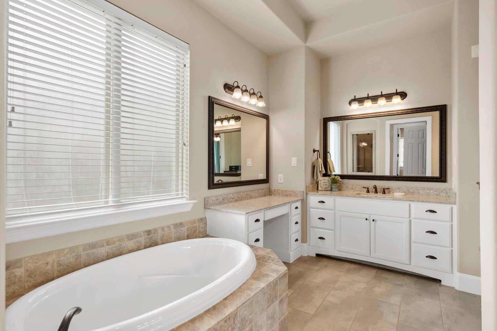 34 Stefano Way, Missouri City, TX 77459 Primary bathroom view showcasing a deep oval soaking tub set in a custom tile surround beneath a large privacy-glass window, alongside a built-in makeup vanity area with granite counter and drawers.