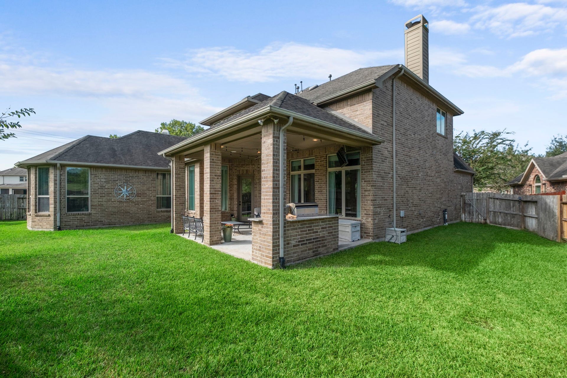 34 Stefano Way, Missouri City, TX 77459 Full rear view of the two-story brick home, showcasing the integrated covered patio, outdoor kitchen structure, stone chimney stack, and gabled roofline.