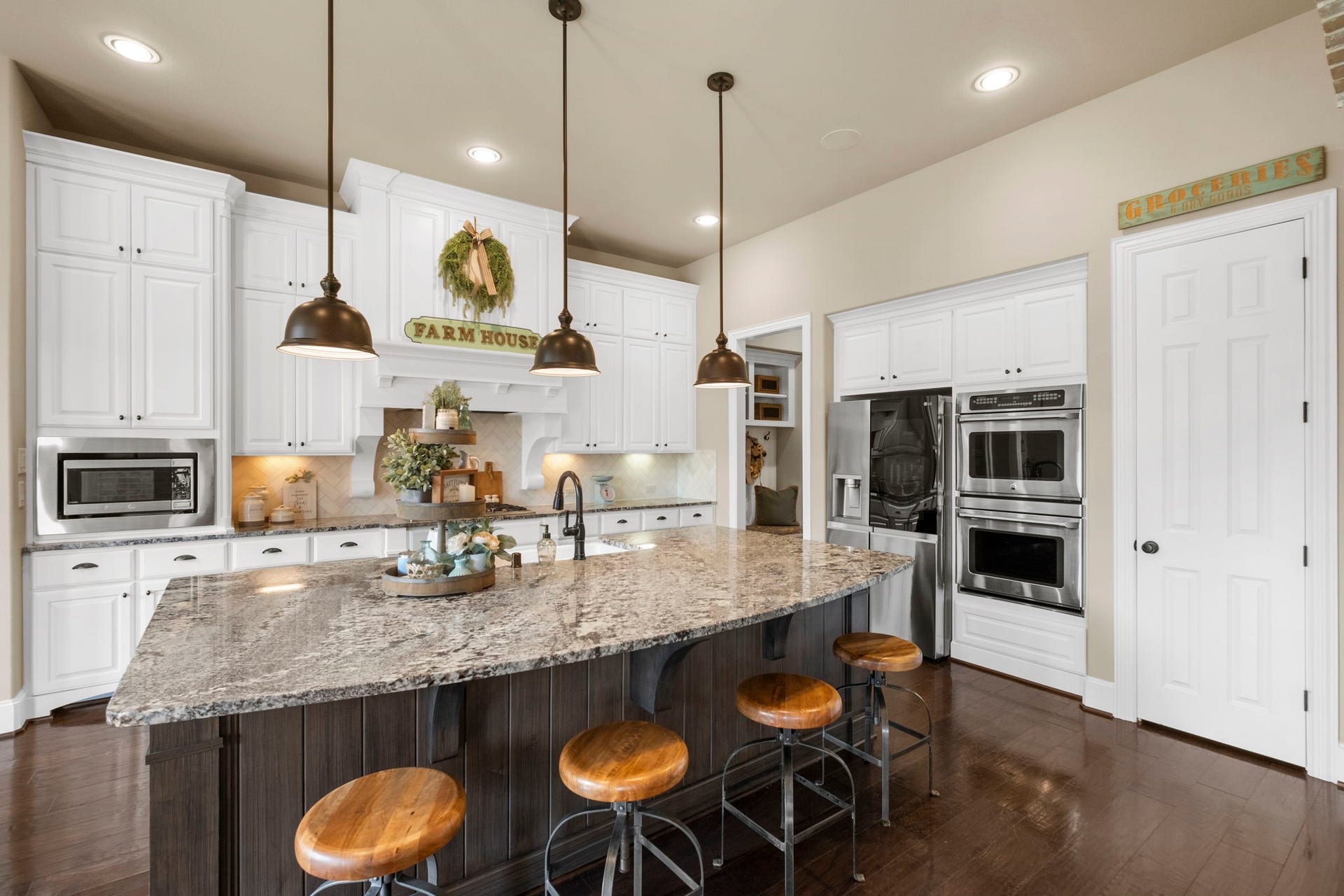 34 Stefano Way, Missouri City, TX 77459 Functional kitchen perspective showing double wall ovens, a built-in microwave surround, integrated stainless steel appliances, and a walk-in pantry door. Highlights the extensive granite counter space and raised panel cabinet layout.