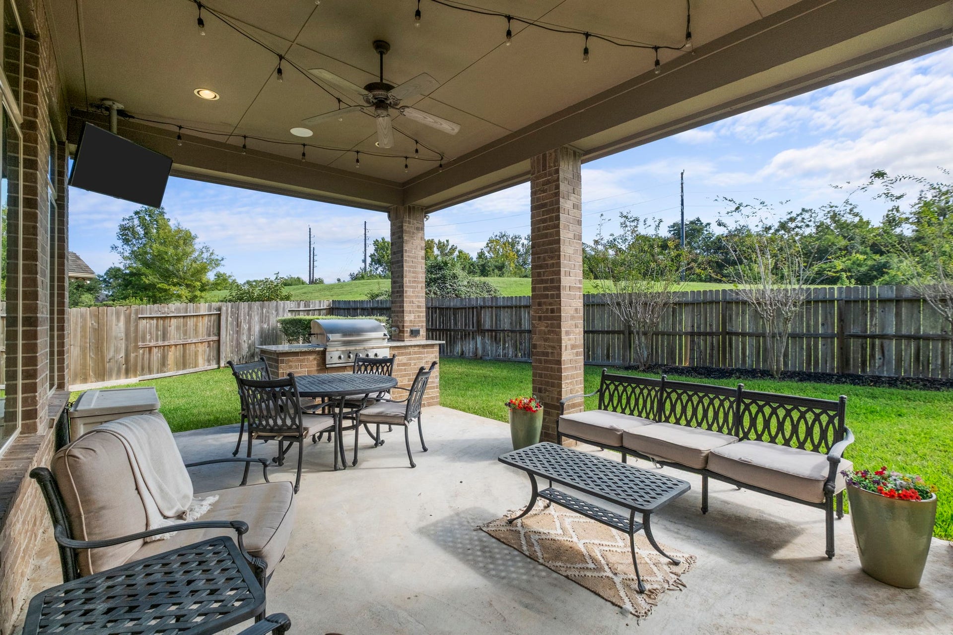 34 Stefano Way, Missouri City, TX 77459 Spacious covered back patio featuring a beadboard ceiling with a ceiling fan, recessed lighting, and outdoor string lights. Equipped with a built-in outdoor kitchen with granite countertops, integrated grill, outdoor TV mount, and stone support pillars.