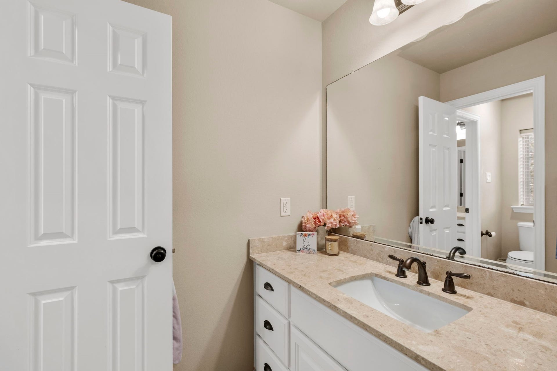 34 Stefano Way, Missouri City, TX 77459 Detail view of the second-story bathroom vanity space showcasing white raised-panel cabinetry, dark drawer pulls, oil-rubbed bronze plumbing fixtures, neutral wall finish, and a wide plate-glass vanity mirror reflecting light from the adjacent hallway and bedroom.