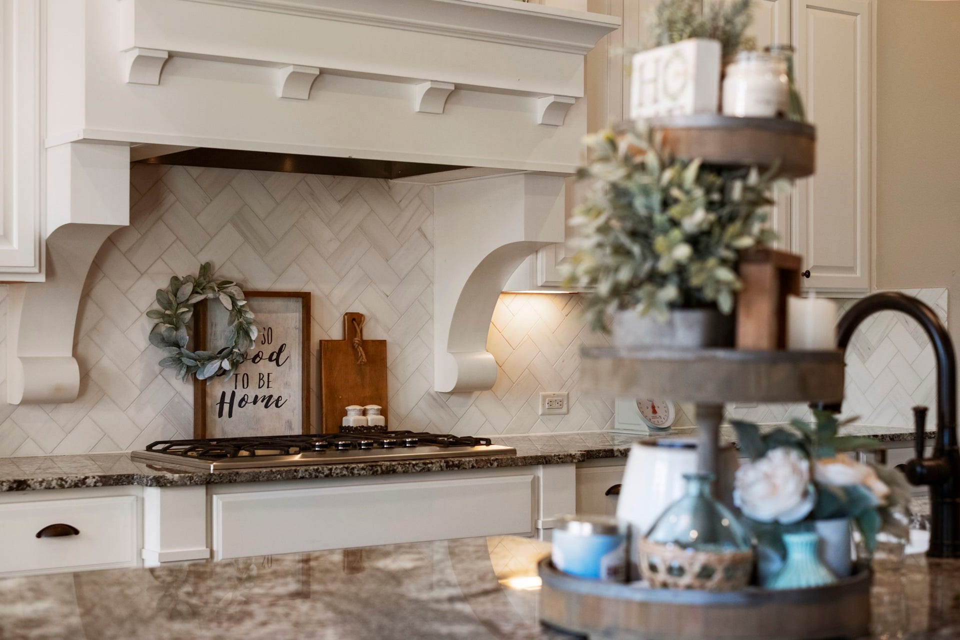 34 Stefano Way, Missouri City, TX 77459 Detailed view of the custom white-painted wood range hood featuring carved corbels, built over a gas cooktop set in granite counter space. Highlights the light-toned subway tile arranged in a classic herringbone backsplash pattern with under-cabinet accent lighting.