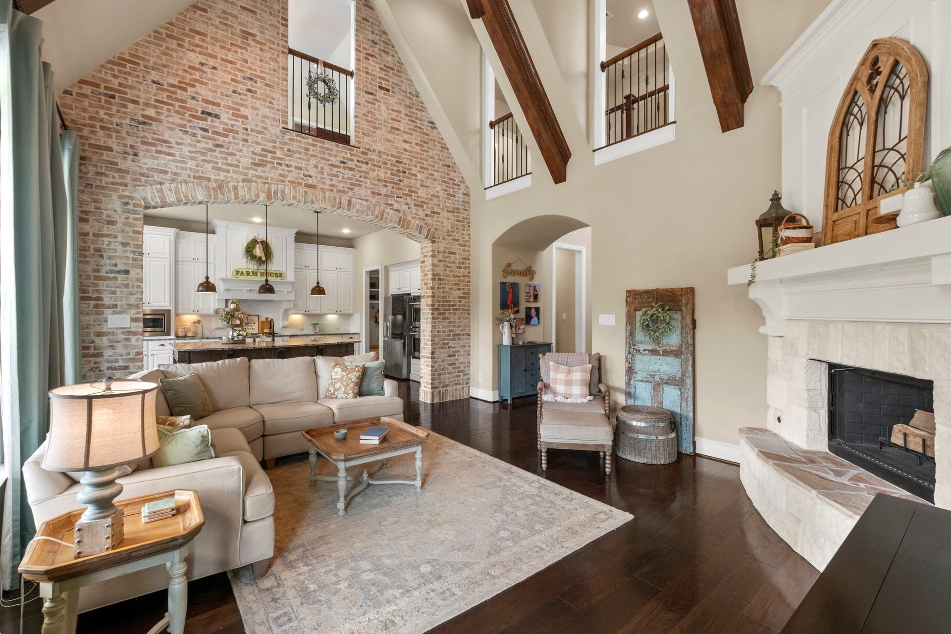 34 Stefano Way, Missouri City, TX 77459 High-angle living room perspective showcasing the full double-height ceiling, upper-level hallway overlooks with wrought-iron railings, exposed wood beams, and a full accent wall crafted in exposed brick with an oversized architectural archway.