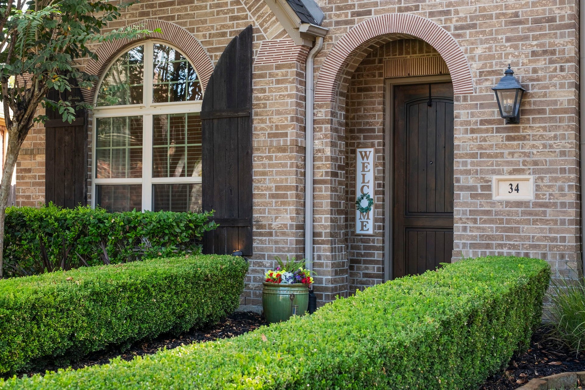 34 Stefano Way, Missouri City, TX 77459 Close-up exterior view highlighting the home's detailed brickwork, double arched entryways with brick header accents, rustic wooden shutters on the multi-pane arched window, a lantern light fixture, and sculpted boxwood hedging.