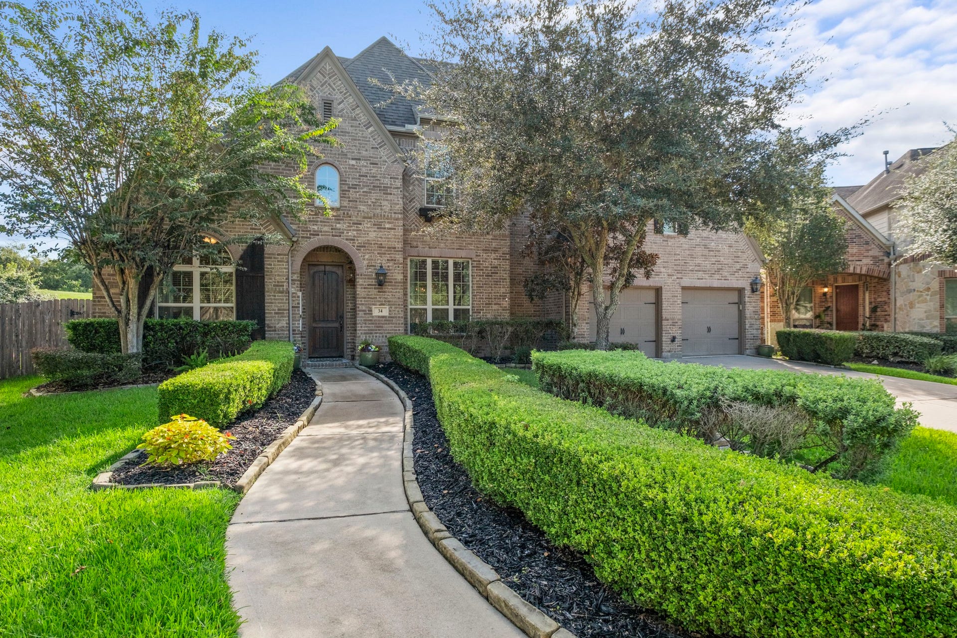 34 Stefano Way, Missouri City, TX 77459 Stately two-story brick residence featuring manicured, stone-bordered landscaping, deep mulch beds, and a winding concrete walkway leading to an arched stone entry. Includes double-pane windows with decorative shutters and a tandem multi-car garage.