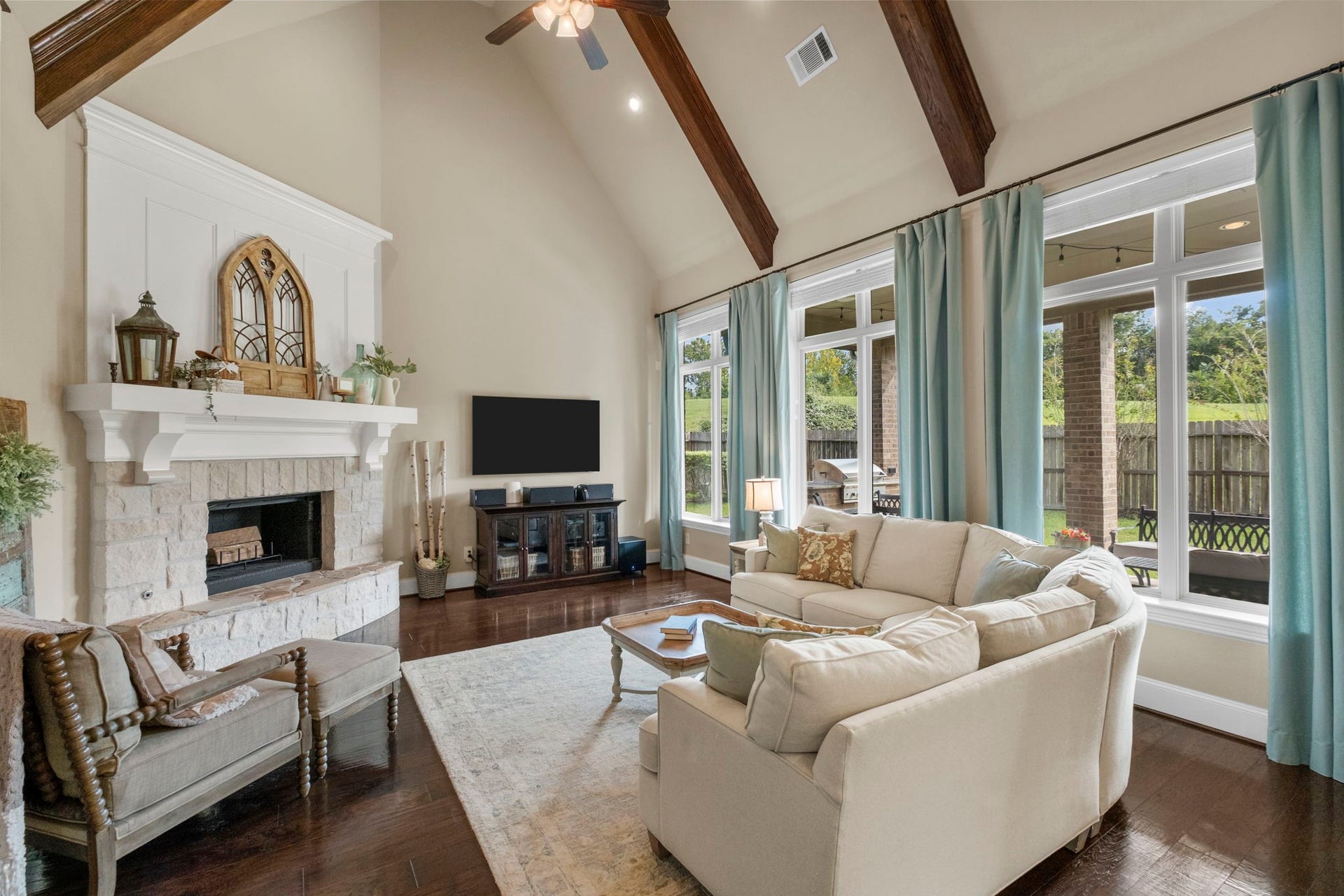 34 Stefano Way, Missouri City, TX 77459 Soaring vaulted ceiling featuring exposed timber beams and large floor-to-ceiling windows overlooking the backyard. The focal point is a floor-to-ceiling floor-mounted stone fireplace with a custom wood mantle and white board-and-batten accent wall above.