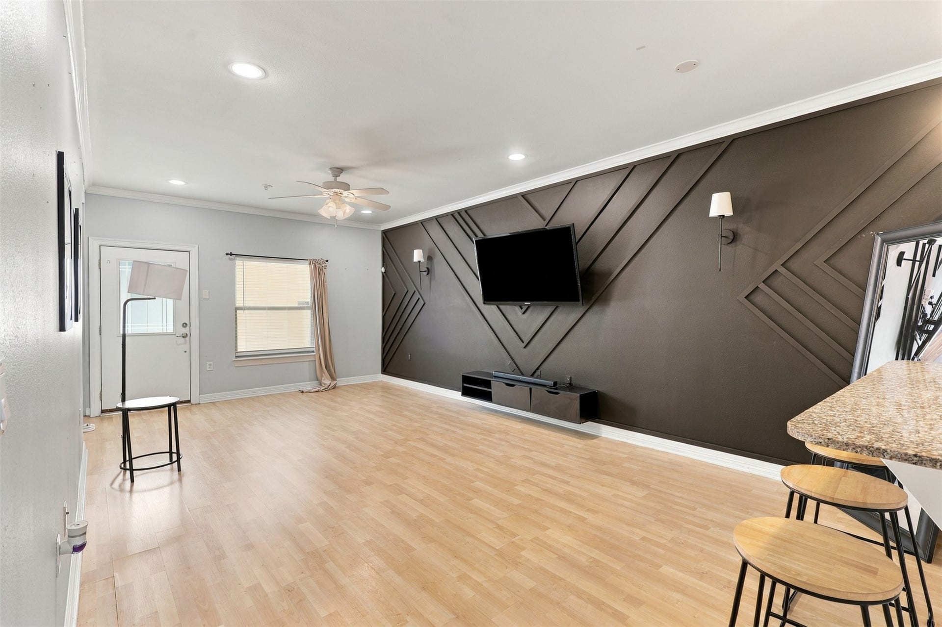 3501 Chenevert Street, #3, Houston, TX 77004 Spacious living area with modern geometric accent wall, featuring a mounted TV and sleek wood flooring. Ample natural light flows through the window, complemented by recessed lighting and a ceiling fan. A granite breakfast bar with stool seating adds a touch of elegance.