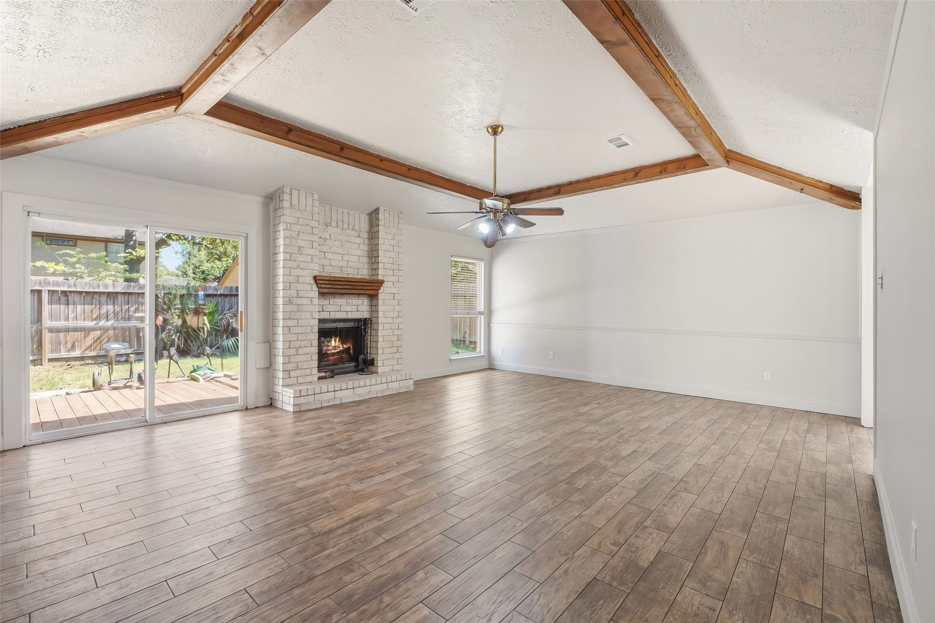 25806 Old Carriage Lane, Spring, TX 77373 Spacious living room with vaulted ceilings and wood beams, featuring a charming white brick fireplace. Abundant natural light pours in through large windows and sliding glass doors, offering easy access to the backyard.