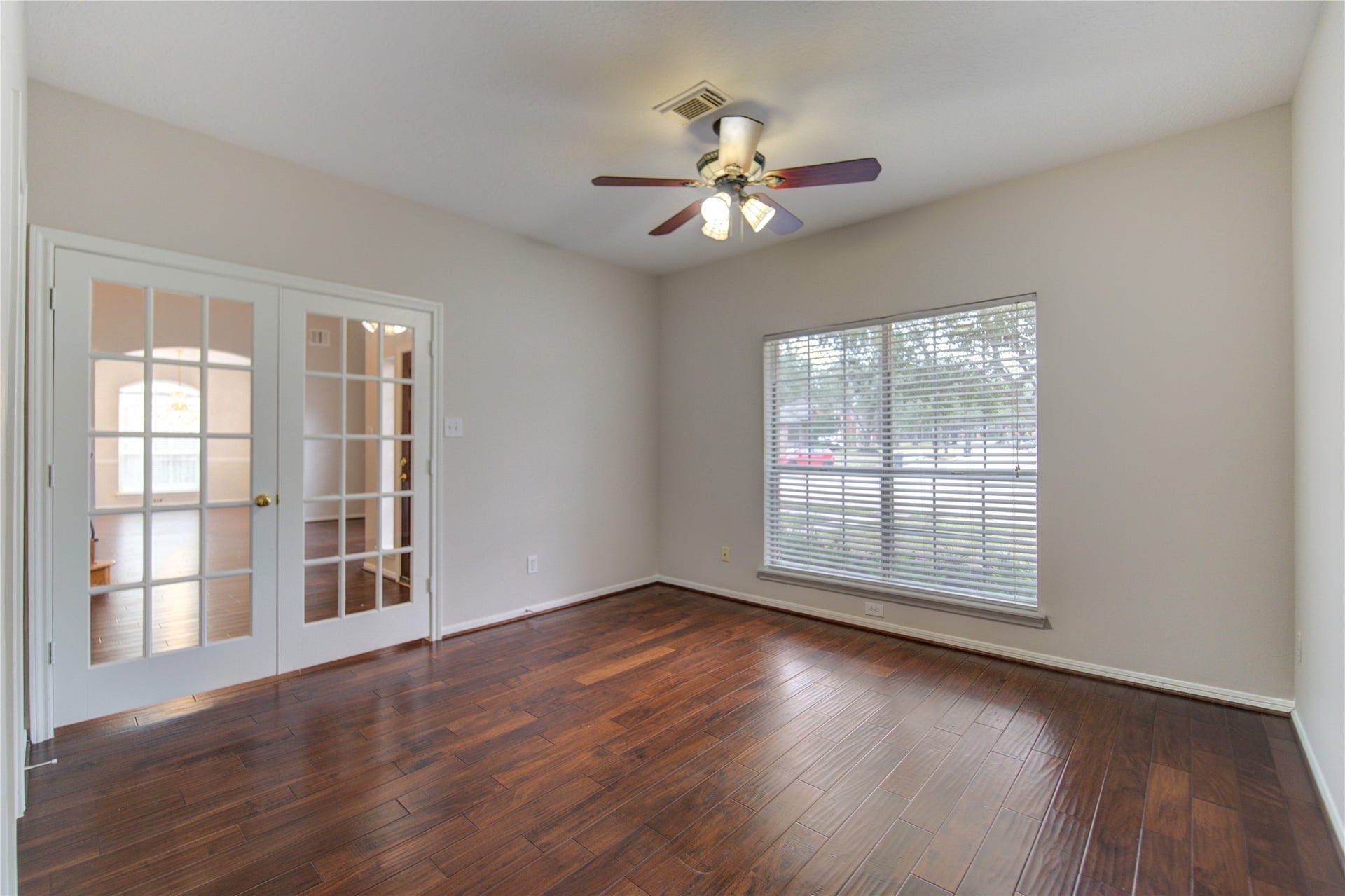 Bright, spacious room with large window, hardwood floors, neutral walls, and a ceiling fan, perfect for a bedroom or office.