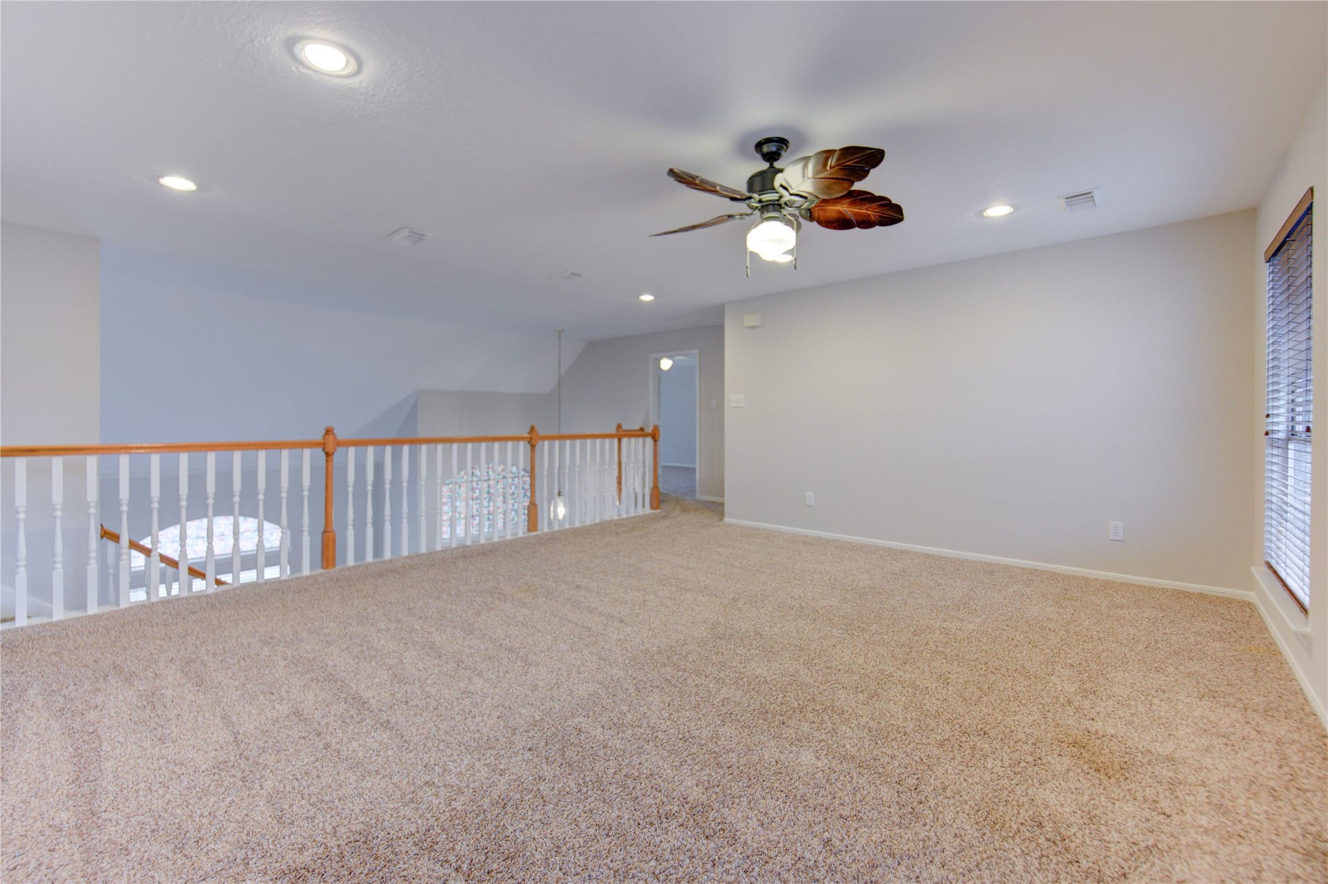 Spacious, carpeted game room with neutral walls, recessed lighting, ceiling fan, two large windows with blinds, and modern wall art, perfect for relaxation or entertaining.