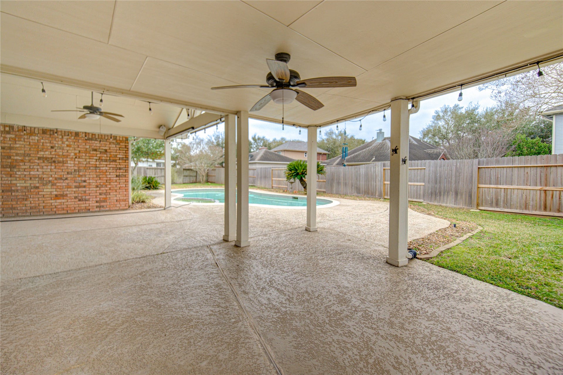This spacious backyard features a charming two-story brick and siding house with a covered patio ideal for outdoor entertaining, and a beautiful in-ground pool perfect for relaxation and summer fun. The surrounding area is fenced for privacy, with some greenery enhancing the serene atmosphere.