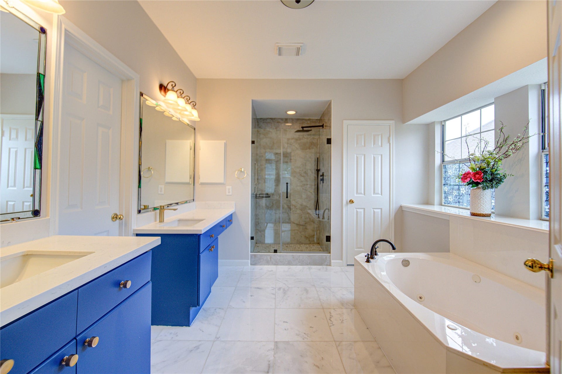 This spacious bathroom features a soaking tub and new innovated shower room, dual blue vanities with ample counter space, large mirrors, and elegant lighting. The bright, open layout is complemented by marble flooring and a window for natural light.