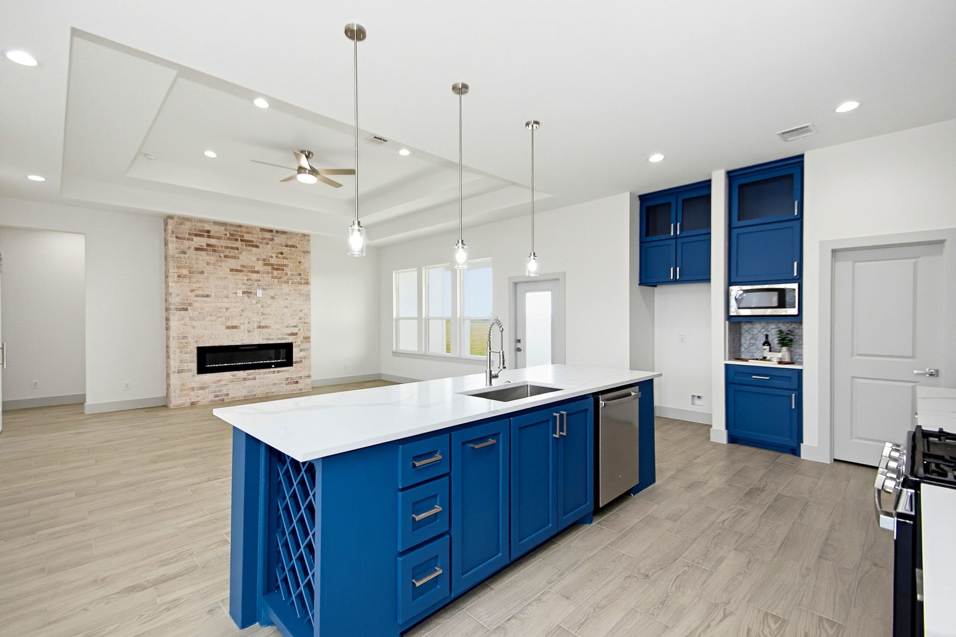 619 Longhorn Trail, Angleton, TX 77515 Modern 19x14 kitchen with striking blue cabinetry, a spacious island with a built-in wine rack, and elegant pendant lighting. The open-concept living area features a cozy brick fireplace and sleek flooring, enhancing the contemporary ambiance.