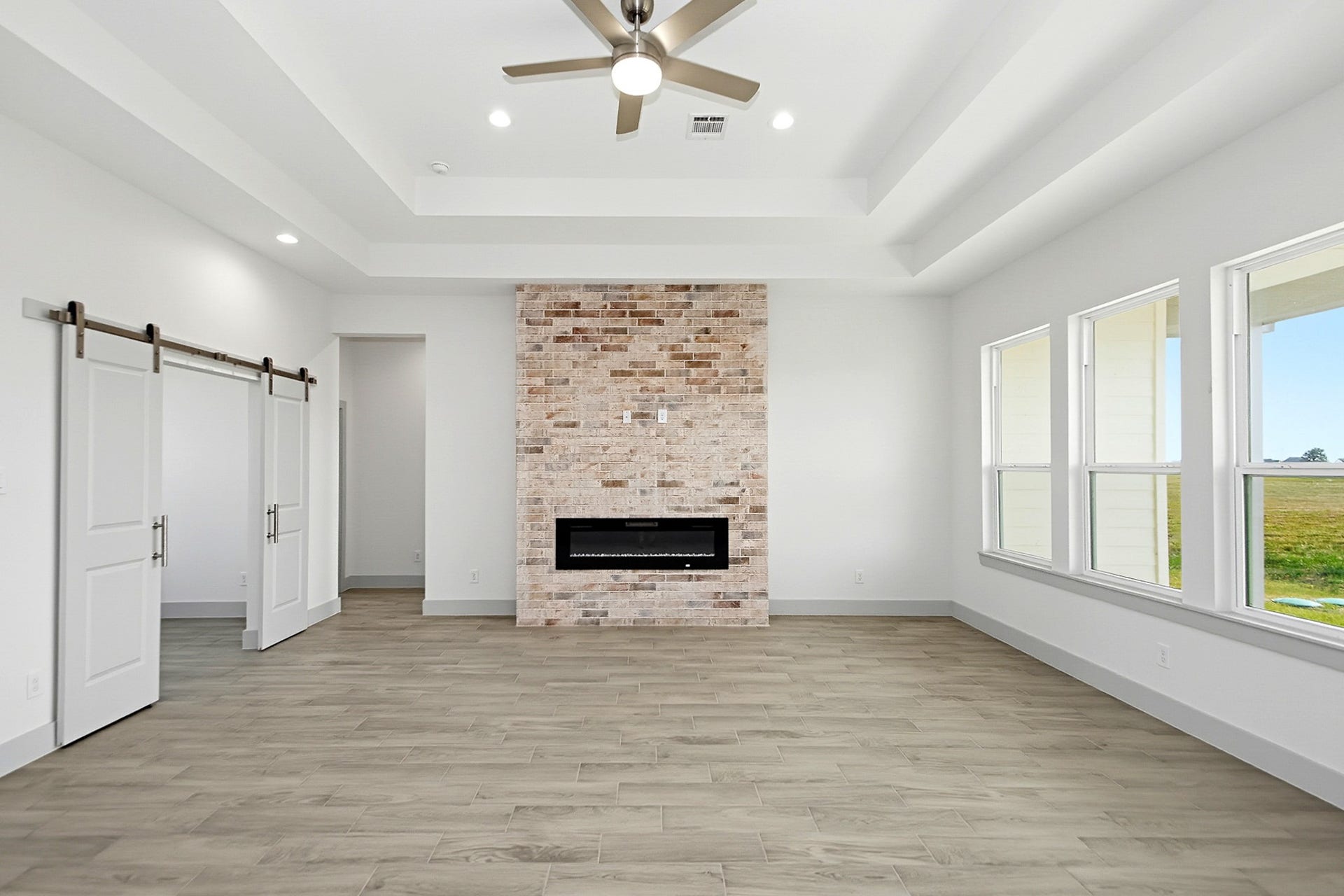 619 Longhorn Trail, Angleton, TX 77515 Spacious living area featuring a modern brick fireplace, tray ceiling with recessed lighting, and large windows that fill the room with natural light. Sliding barn doors add a stylish touch to this office space or extra bedroom if needed.