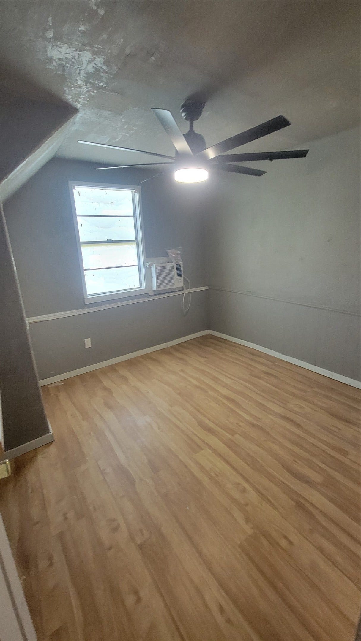 12018 Sandpiper Road, Angleton, TX 77515 This room features laminate wood flooring, a ceiling fan with light, a window with an air conditioning unit, and neutral gray walls. It offers a cozy space with a sloped ceiling, making it ideal for a small bedroom or office.