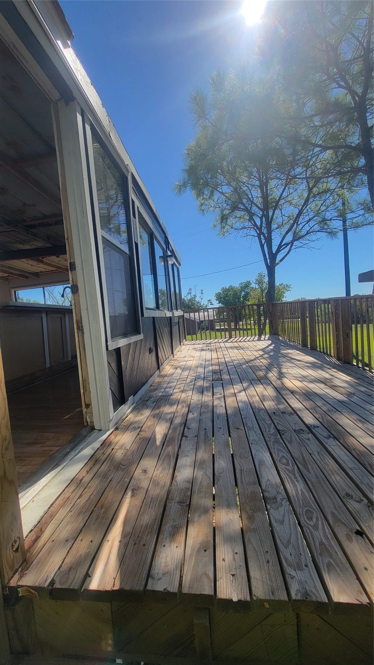 12018 Sandpiper Road, Angleton, TX 77515 This photo shows a wooden deck attached to a house, partially covered by a porch with large windows. The deck is spacious and surrounded by a railing, with a tree nearby providing some shade. The area looks sunny and inviting.