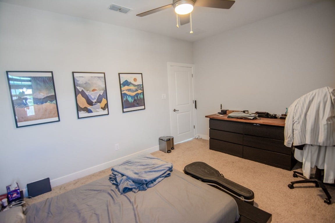 5423 Priamus Drive, Missouri City, TX 77459 This bedroom features neutral walls, a ceiling fan, and carpeting. It has a simple layout with wall art, a bed, a dark dresser, and a desk chair. There's a door leading to another area, providing a cozy and functional space.