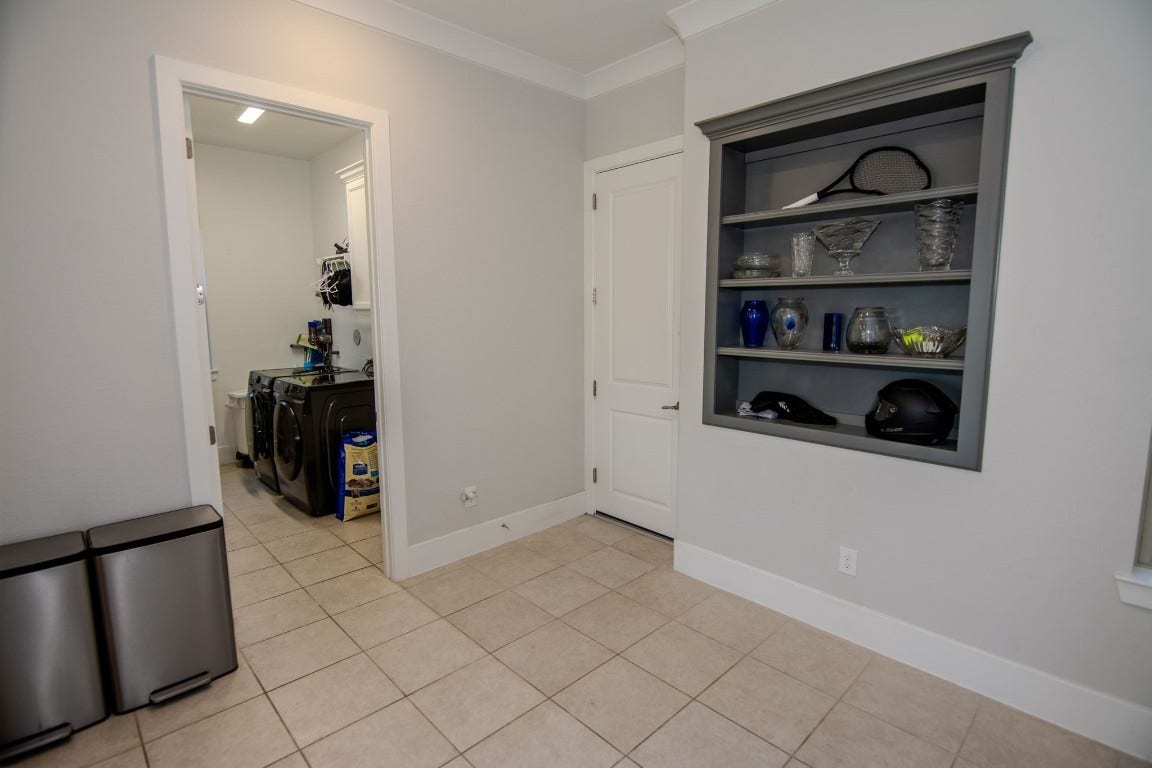 5423 Priamus Drive, Missouri City, TX 77459 This photo showcases a clean, tiled utility space featuring a laundry area with modern appliances. There's a built-in shelf displaying decorative items, and a storage closet is visible. The room is well-lit and functional.