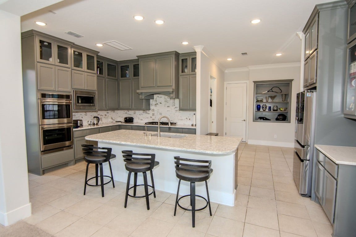 5423 Priamus Drive, Missouri City, TX 77459 This photo showcases a modern kitchen with gray cabinetry, stainless steel appliances, and a large central island with seating for three. The space is well-lit with recessed lighting and features a stylish tile backsplash.