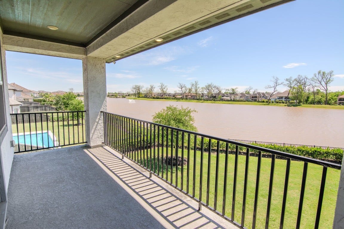 5423 Priamus Drive, Missouri City, TX 77459 This photo showcases a spacious covered balcony with a black railing, offering a serene view of a large, calm lake. The surrounding greenery and glimpses of neighboring homes enhance the tranquil setting, perfect for relaxation and enjoying the outdoors.